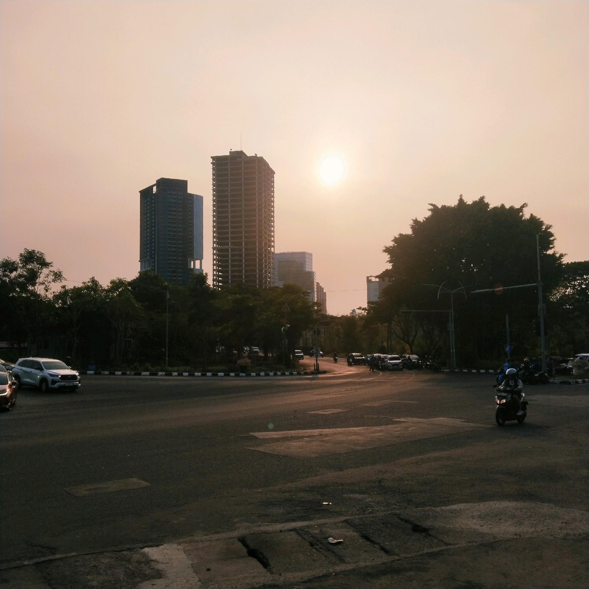 This image depicts a street scene in Surabaya, Indonesia, during what appears to be late afternoon or early evening, indicated by the low sun casting a warm, diffused light. The sky is hazy, with the sun visible as a bright, circular orb. In the background, several tall buildings, including a partially constructed skyscraper, rise above a dense line of trees. The foreground features a road intersection with several vehicles, including cars and motorcycles. A lone motorcyclist, wearing a helmet, is in motion on the right side of the frame. Traffic appears to be moderate, with vehicles on various lanes of the road. The overall environment suggests an urban setting with ongoing development.