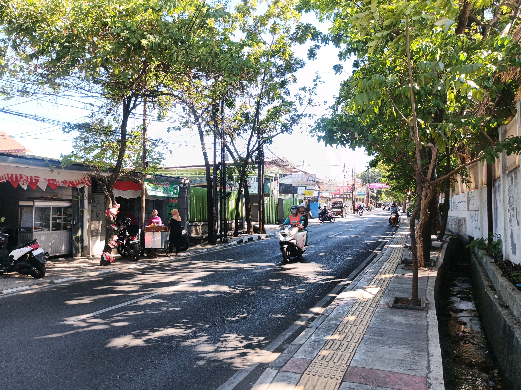 This is a street-level view of a bustling road in Bandung City, Indonesia, captured under a bright, sunny sky. The road is paved, with a clear white lane marking down the center. To the left, a row of modest buildings lines the street, some featuring awnings and decorated with red and white banners, suggesting a festive or patriotic occasion. Several motorbikes are parked on the sidewalk in front of these buildings.
On the right side of the road, a paved sidewalk is visible, bordered by a shallow concrete drainage ditch that contains murky water and debris. Trees with lush green foliage are planted along the sidewalk, casting dappled shadows onto the road and pedestrian area.
Several people are present. On the left side, near the buildings, two women are interacting with a street vendor selling food from a cart. Further down the street, two individuals are riding a motorbike, and another motorbike is visible approaching from the distance. The overall atmosphere is one of everyday urban activity.