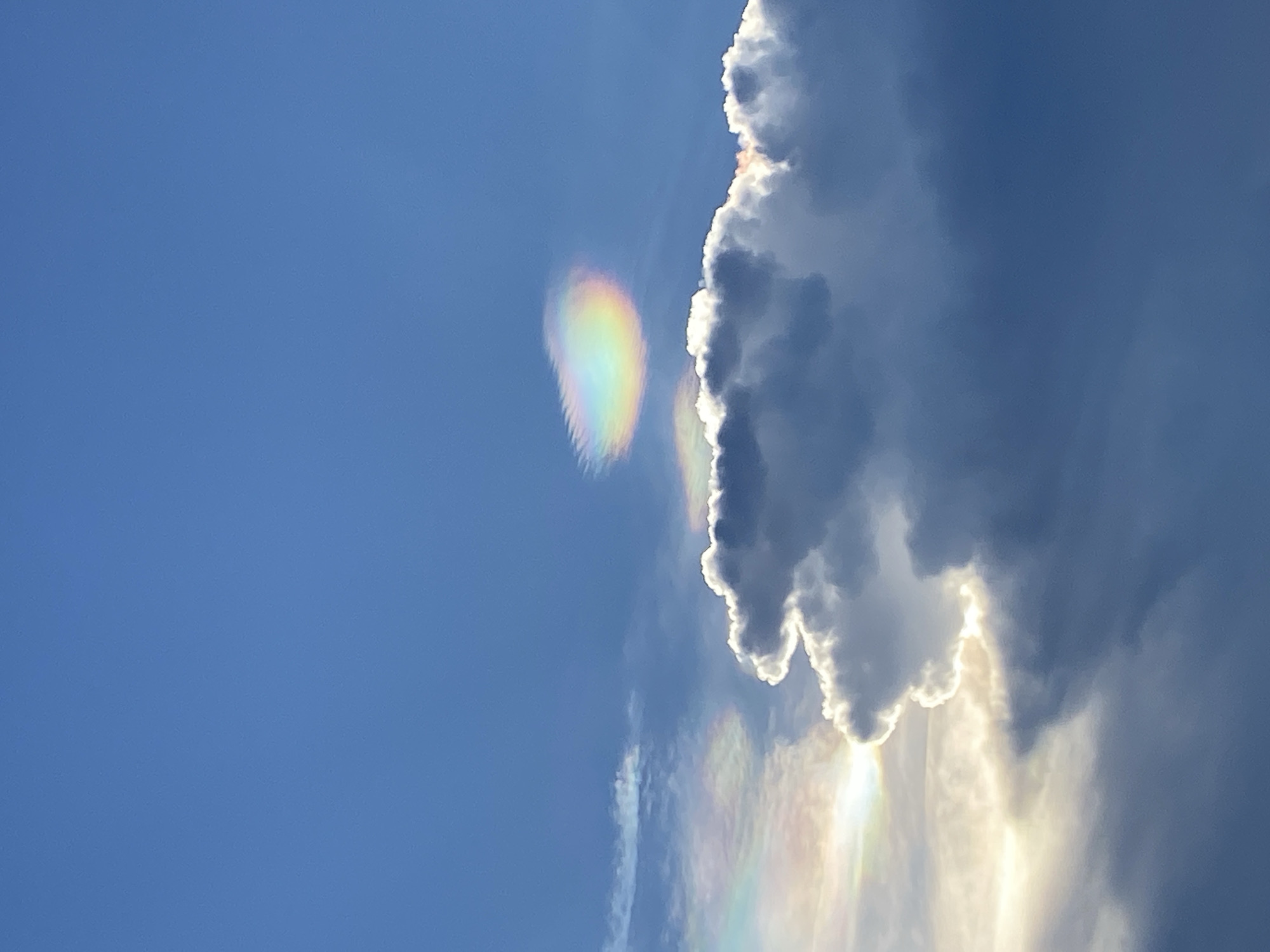 This is a vertical, eye-level shot of a partly cloudy sky over Mui Ne, Vietnam, with a focus on iridescence in the clouds. The sky is a deep, clear blue in the upper portion of the frame. In the middle and lower portions of the image, large, dark gray clouds dominate, with their edges brightly illuminated by the sun, creating a stark contrast. Above these darker clouds, and to the left and right, are thinner, wispy clouds. The most striking feature is the presence of cloud iridescence, visible as a rainbow-like sheen on some of the smaller, higher clouds and also along the illuminated edges of the larger clouds. One particularly prominent iridescent cloud formation is a rounded, elongated shape in the upper middle of the frame, displaying vibrant bands of red, orange, yellow, green, and blue. The overall impression is a dynamic sky with dramatic lighting and atmospheric optical phenomena.
