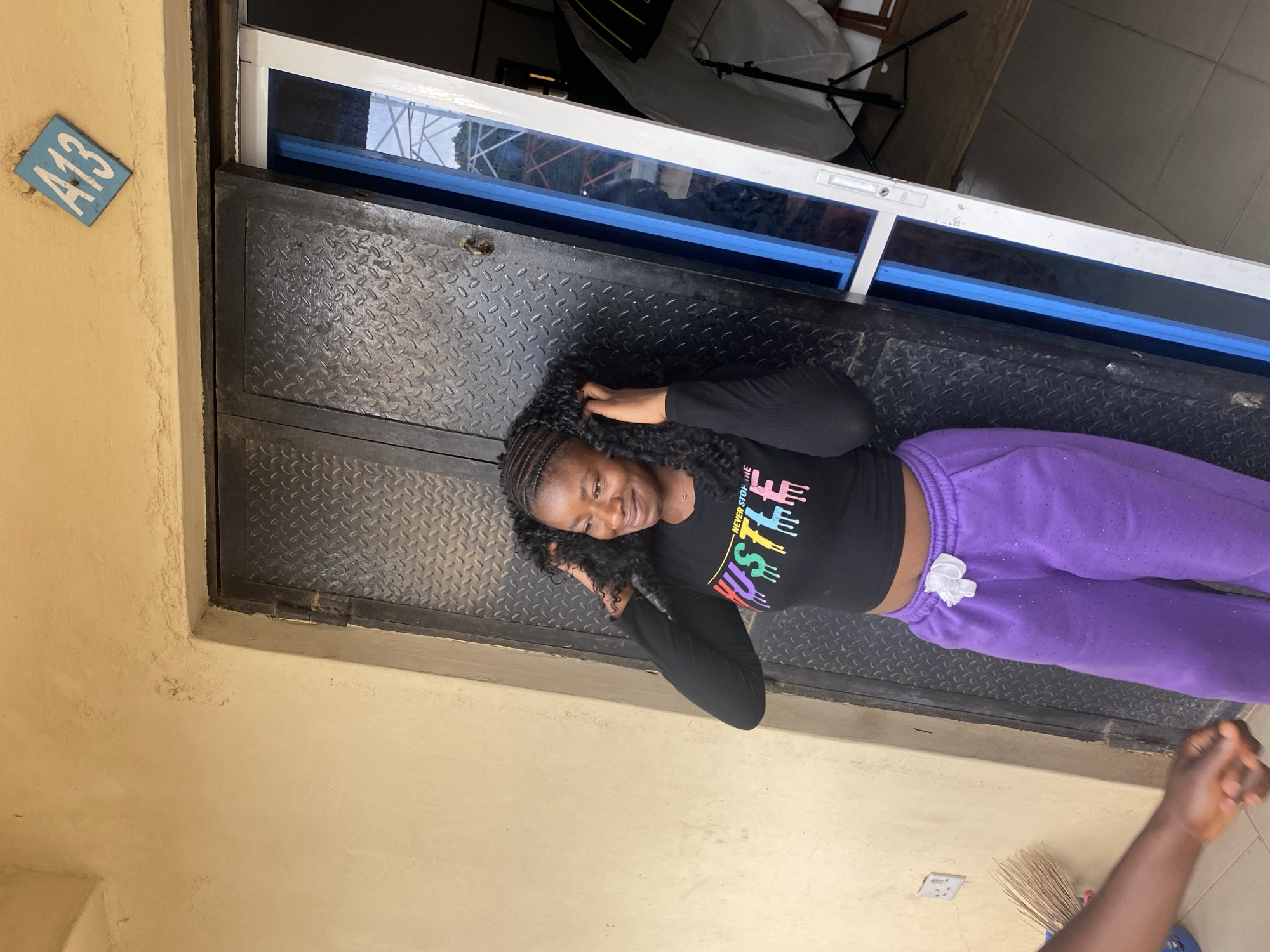 A young Black woman is depicted lying horizontally on a dark, textured surface, possibly a metal platform or door, with a raised diamond pattern. She has braided hair and is smiling warmly, looking towards the camera with her hands near her head. She is dressed in a black long-sleeved crop top featuring colorful, dripping letters that spell "HUSTLE" above which "NEVER STOP THE" is subtly visible. Her lower body is clad in sparkly purple sweatpants.
Above the woman's head, there is a white-framed window or display case with a blue tinted pane, reflecting what appears to be outdoor elements and some photographic studio equipment, including a softbox and a light stand, in the background. To the left of the woman and the dark surface, a light yellow-beige wall is prominent. A small, light blue rectangular sign with "A13" written in white is affixed to this wall. In the lower right corner of the image, a dark-skinned arm and hand extend into the frame. Partially visible below the arm on the yellow wall are a brown broom and a white electrical outlet. The setting appears to be an indoor or semi-enclosed area, likely during the day in Bukuru, Nigeria, with the woman exuding a relaxed and cheerful demeanor.