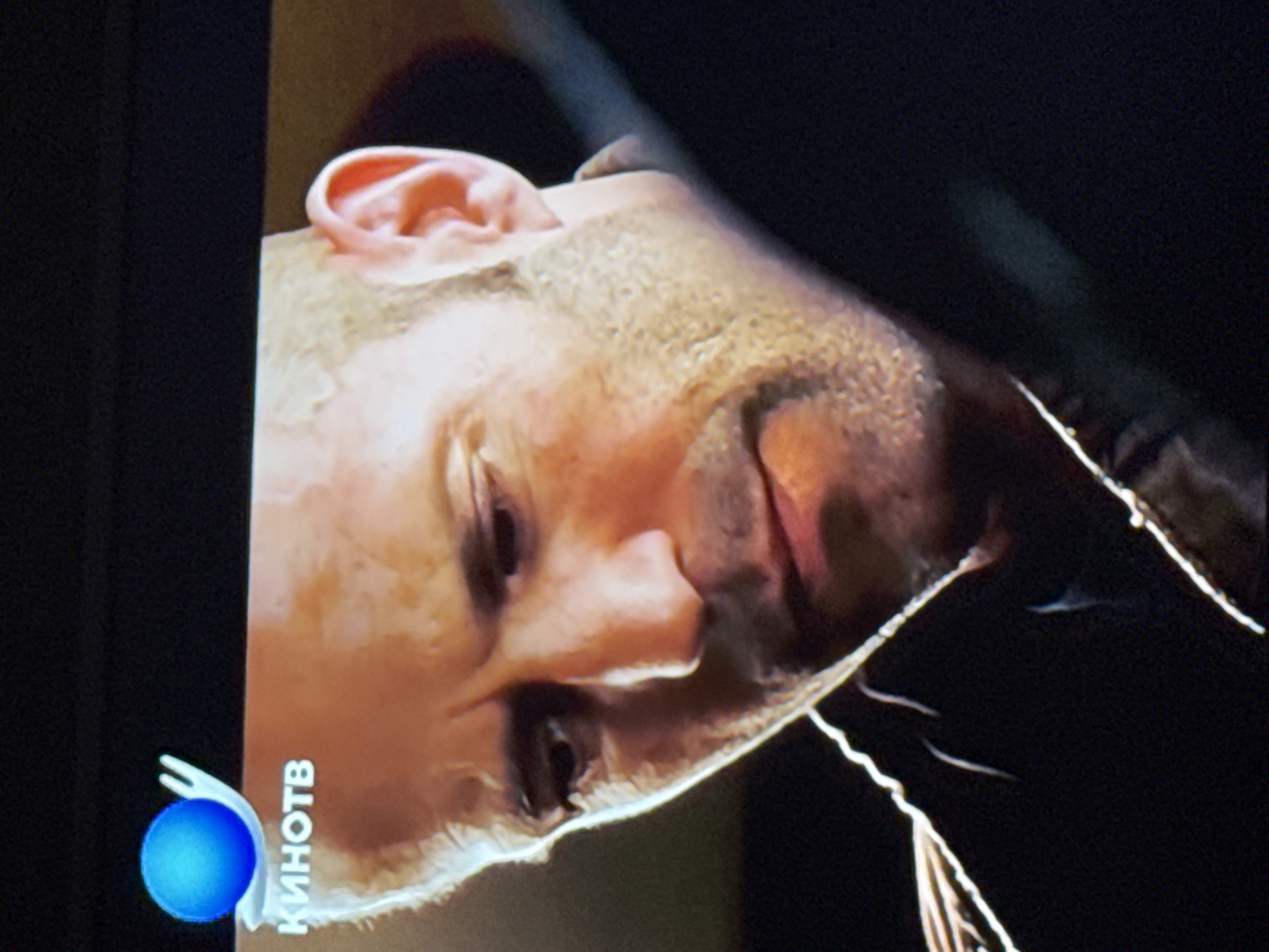 The image is a photograph of a television screen displaying a man's face. The man has a shaved head and light skin. He appears to be looking down with his eyes partially closed. The television channel's logo "КиноТВ" is visible in the bottom left corner of the screen. The background is dark. The location is not discernible from the image.