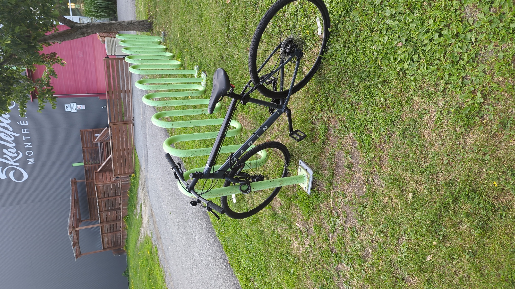 A dark gray Marin bicycle is parked in a green metal bike rack in front of a grey building with "SKATEPARK MONTRÉAL" written in large white script. The building has a large pink section to the right and a wooden structure with a roof and railings to the left. The bike is locked to the rack with a black U-lock and a cable lock. The bike has a front fender, a rear rack, and is a hybrid or commuter style bicycle. The ground is covered in green grass, and there is a paved path behind the bike rack. The environment appears to be an outdoor public space.