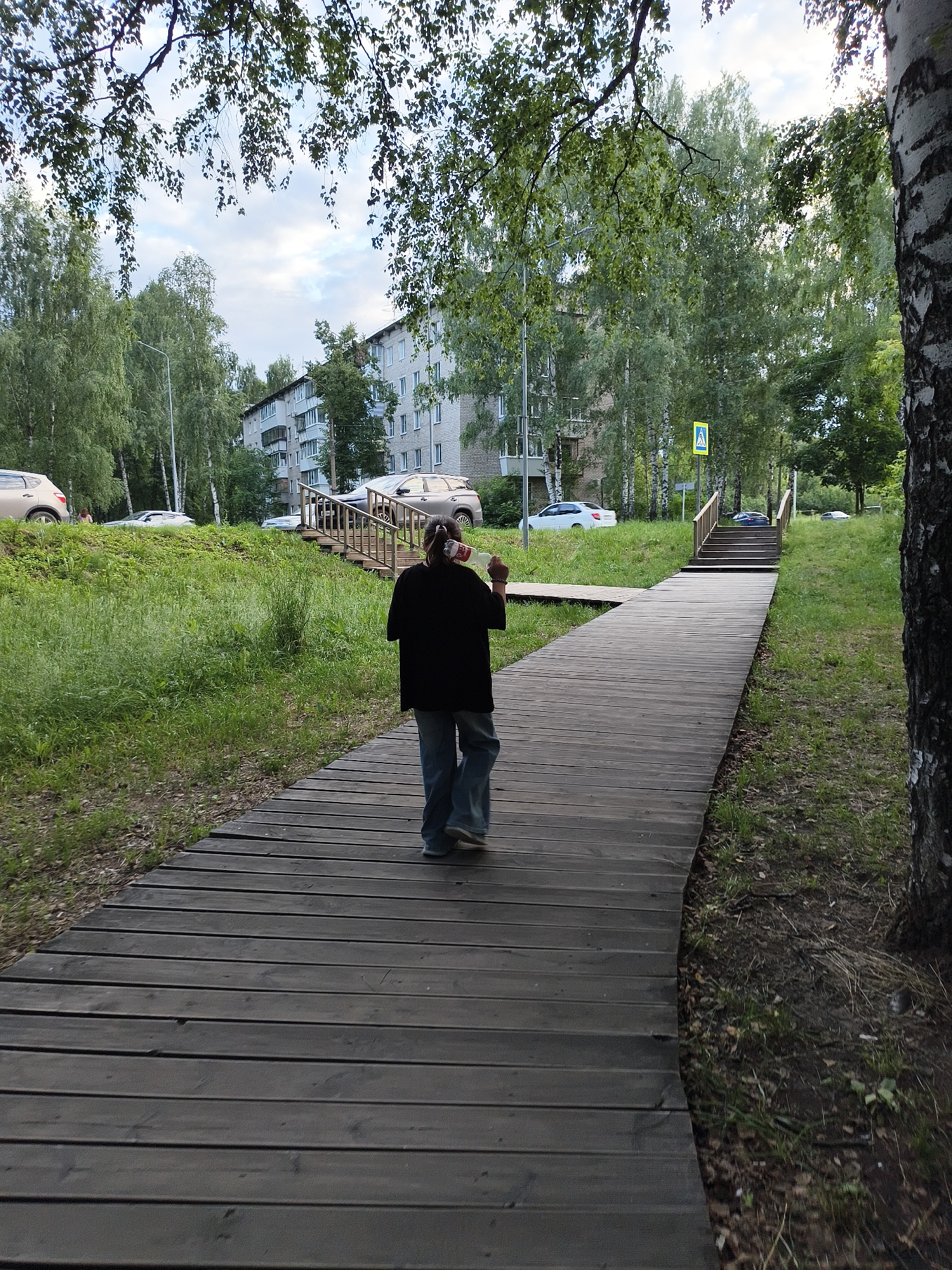 In Chaikovsky, Russia, a person with their back to the camera walks along a dark wooden boardwalk. The person is wearing a black jacket and blue jeans. The boardwalk curves to the right and appears to be made of planks laid horizontally. To the left of the boardwalk is a grassy slope with scattered leaves. In the background, there are trees, a multi-story building, and parked cars. Further down the boardwalk, there is a wooden staircase leading upwards, with a pedestrian crossing sign visible near the top of the stairs. The sky is overcast.