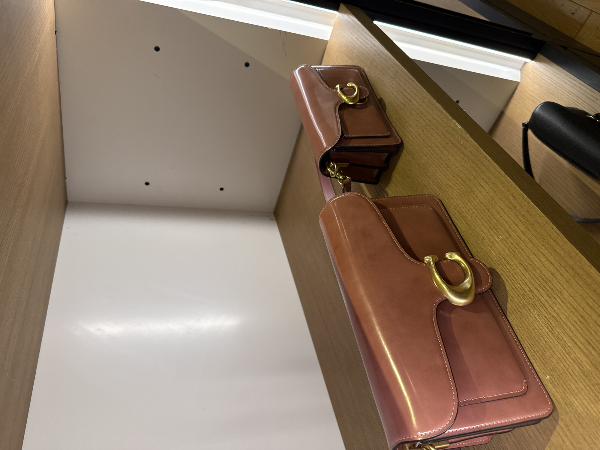 The image shows a display of brown leather purses with gold hardware in a retail setting. The purses are stacked on wooden shelves illuminated by strip lighting. The location is likely within the city of Sunrise, United States of America.