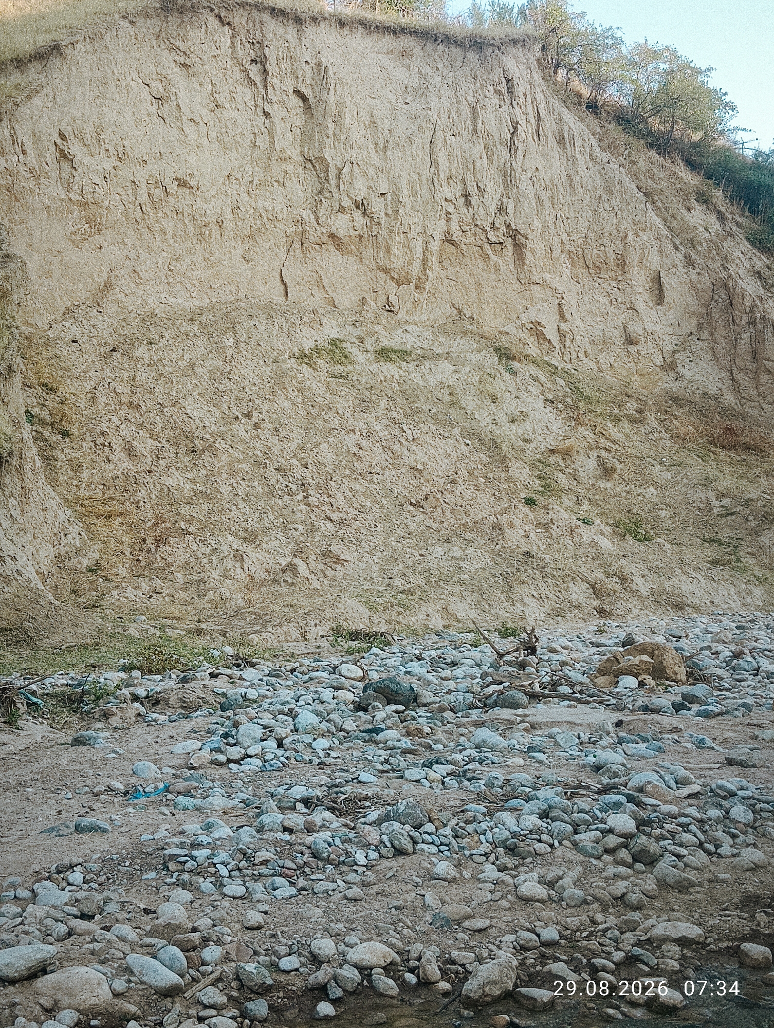 The image shows a steep, eroded cliff face of a riverbank composed of sandy soil and rock. The top of the cliff is covered with sparse vegetation and trees. The riverbed in the foreground is filled with a variety of smooth, rounded stones of different sizes, interspersed with sand and some debris. The scene is outdoors, in a natural environment, and appears to be a dry riverbed or a shallow watercourse. The timestamp "29.08.2026 07:34" is visible in the lower right corner, indicating the date and time the photograph was taken. The location is specified as Ak-Took, Kyrgyzstan. There are no people or animals visible in the image.