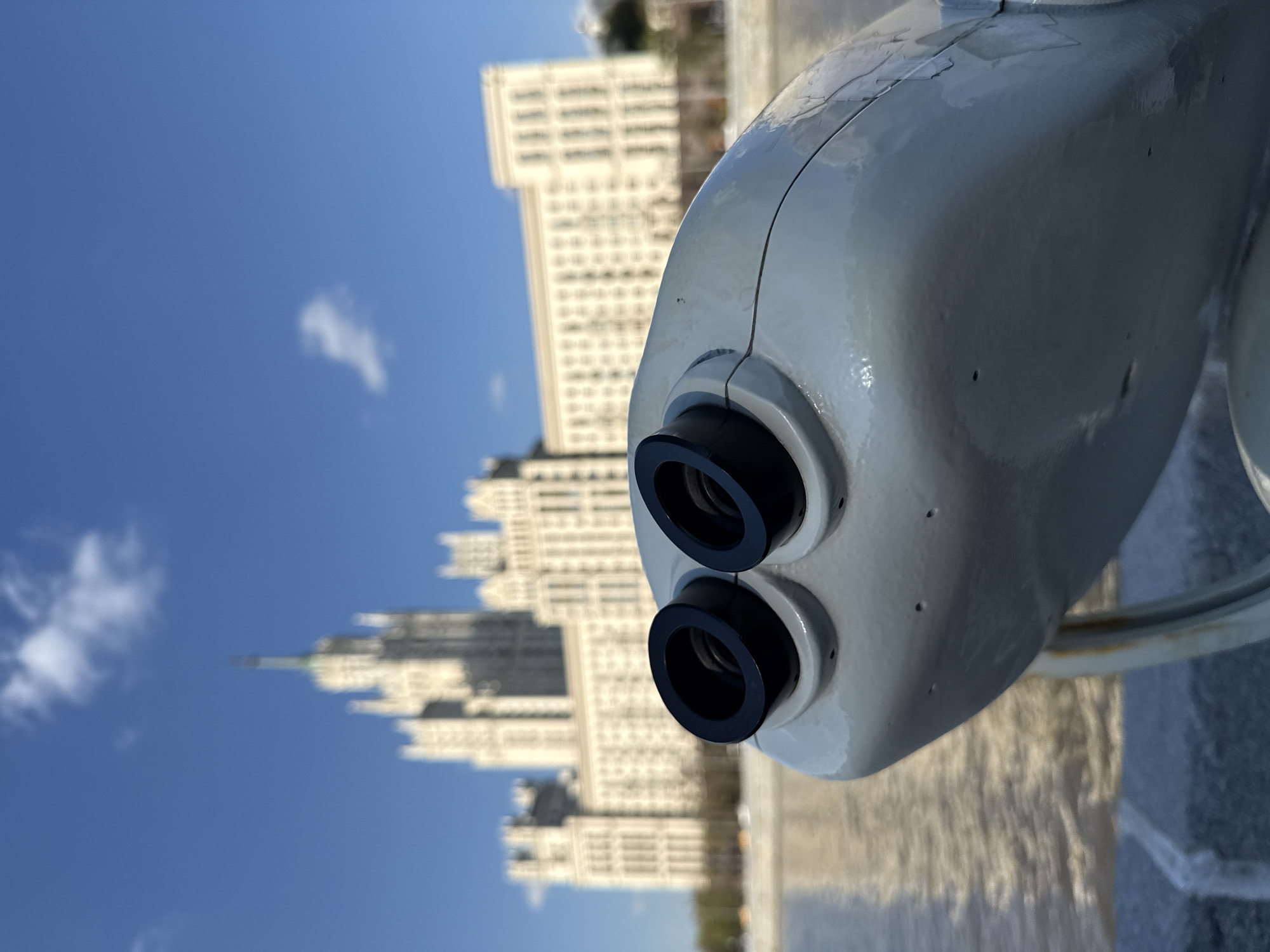 A pair of coin-operated binoculars is in the foreground, angled to the right. Behind the binoculars, in the midground, is one of Moscow’s “Seven Sisters” skyscrapers, likely the Hotel Ukraina. The background shows a clear blue sky with a few scattered clouds. A concrete barrier or wall is visible to the right of the binoculars. The overall scene suggests a tourist location along the Moscow River.