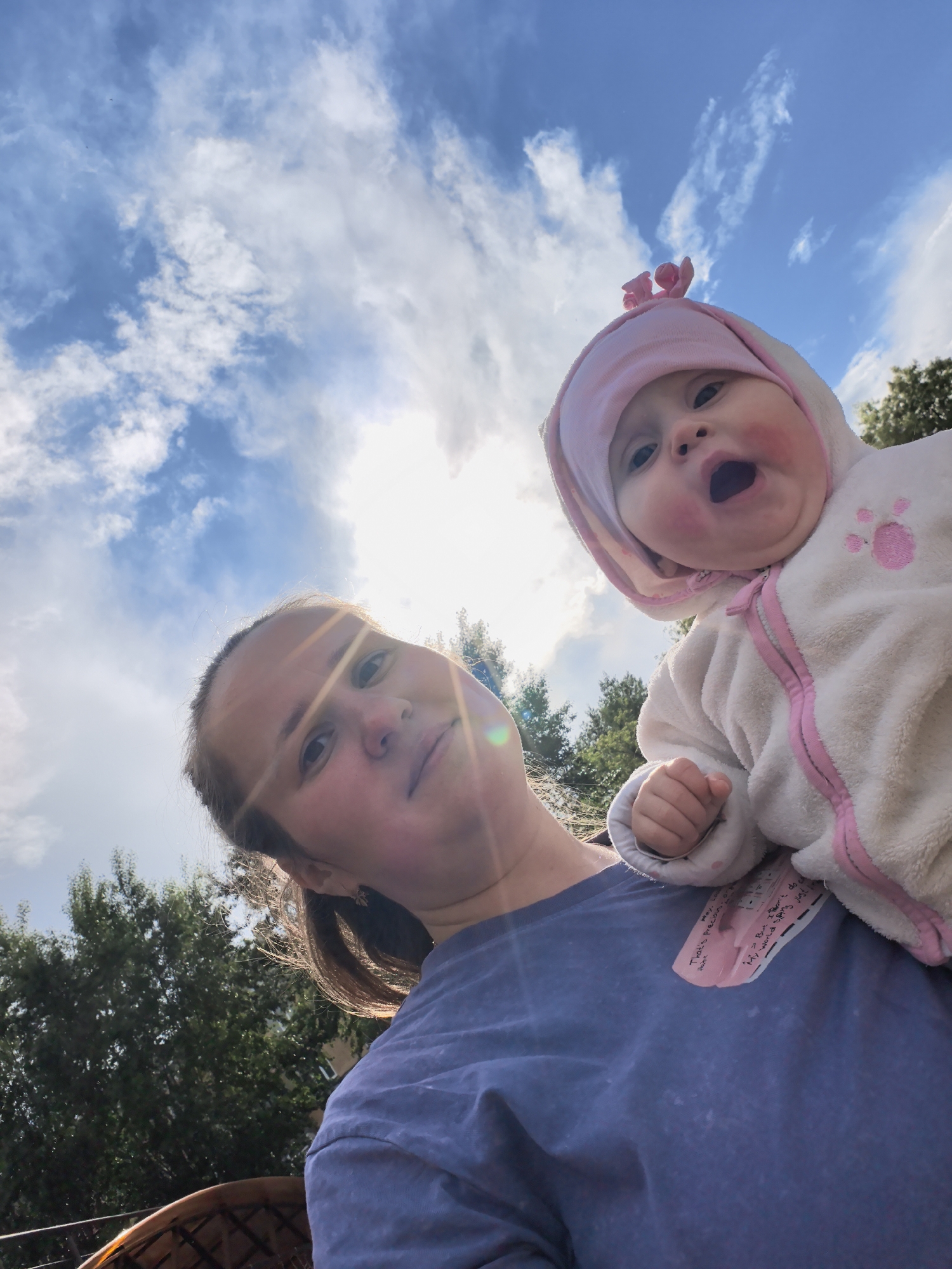 A low-angle medium shot shows a woman holding a baby against a cloudy sky. The woman is wearing a navy blue t-shirt and has her hair pulled back. The baby is dressed in a white, fuzzy hooded snowsuit with pink trim and a pink flower on the hood. The baby's mouth is open. The sky is visible above, with trees in the background. A small note is pinned to the woman's shirt. The location appears to be outdoors, potentially in a park or garden setting, within the city of Unknown, Unknown.