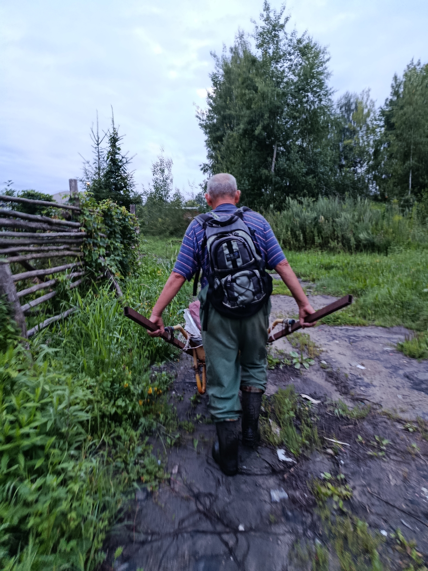 An elderly man with gray hair walks away from the camera, wearing a striped shirt, green pants, and black rubber boots. He carries a backpack and holds a metal frame with a wheel attached, resembling a part of a wheelbarrow or cart, with both hands. He is on a dirt path lined with tall green grass and a wooden fence on the left. Trees and foliage form the background. The scene appears to be in a rural or suburban area on an overcast day.
