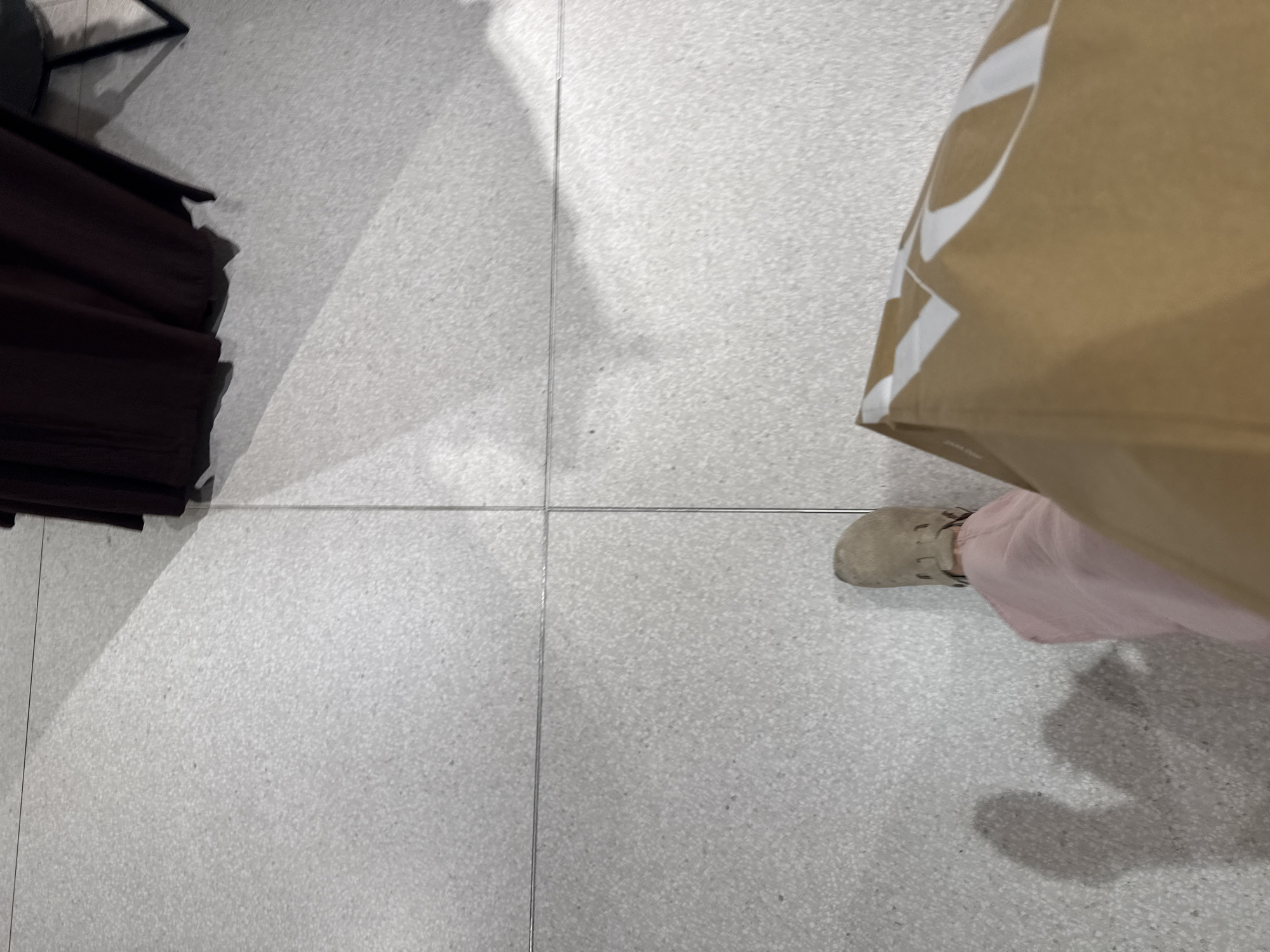 The image shows a first-person perspective of someone standing on light gray tiled flooring. A portion of a dark brown garment rack or clothing is visible in the upper-left corner. A light brown paper shopping bag with white markings is partially in the upper-right corner. The person is wearing a light tan shoe and a light pink garment. The scene appears to be indoors. Without additional information, I can not place the location in the city of Puebla, Mexico.