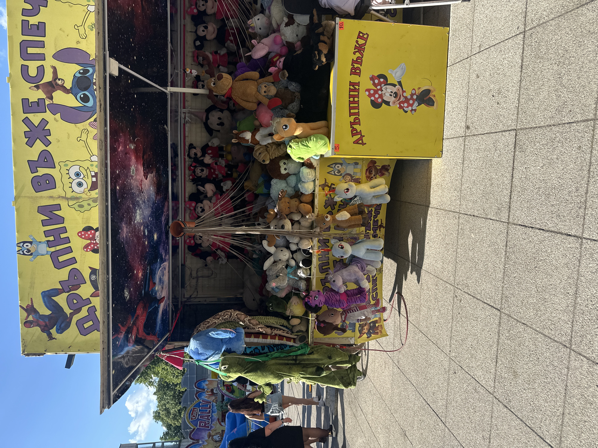 The image captures a prize stall at a fair or market, situated outdoors in daylight. The stall is laden with various stuffed toys hanging from above. Yellow signage bearing images of Mickey Mouse and Spongebob Squarepants flanks the toys. Two individuals are observed in the foreground: one in a black dress and another beside her in shorts. The ground is paved with rectangular tiles. In the background, a tree and signage for "Rally" can be discerned. The location is Montana, Bulgaria.