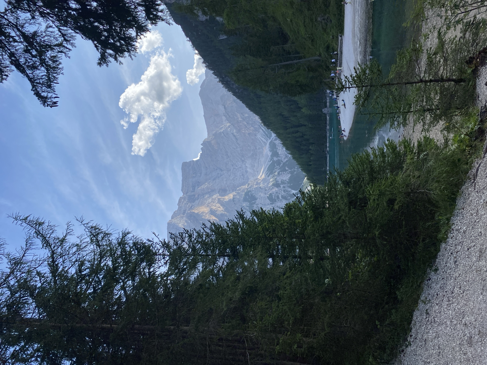 A scenic view of Lago di Braies (Pragser Wildsee) in Prags, South Tyrol, Italy, is captured from a forest trail. In the foreground, a gravel path leads the eye towards a clear, turquoise lake. Dense green pine trees frame the left and right sides of the image, their branches reaching towards the sky. Across the lake, a sandy shore is dotted with numerous people, some walking, some sitting, and a few appear to be sunbathing. Beyond the lake, a vast expanse of dark green forest blankets a steep mountainside. Towering in the background are majestic, rugged mountains, their peaks illuminated by soft sunlight against a blue sky with scattered white clouds.
