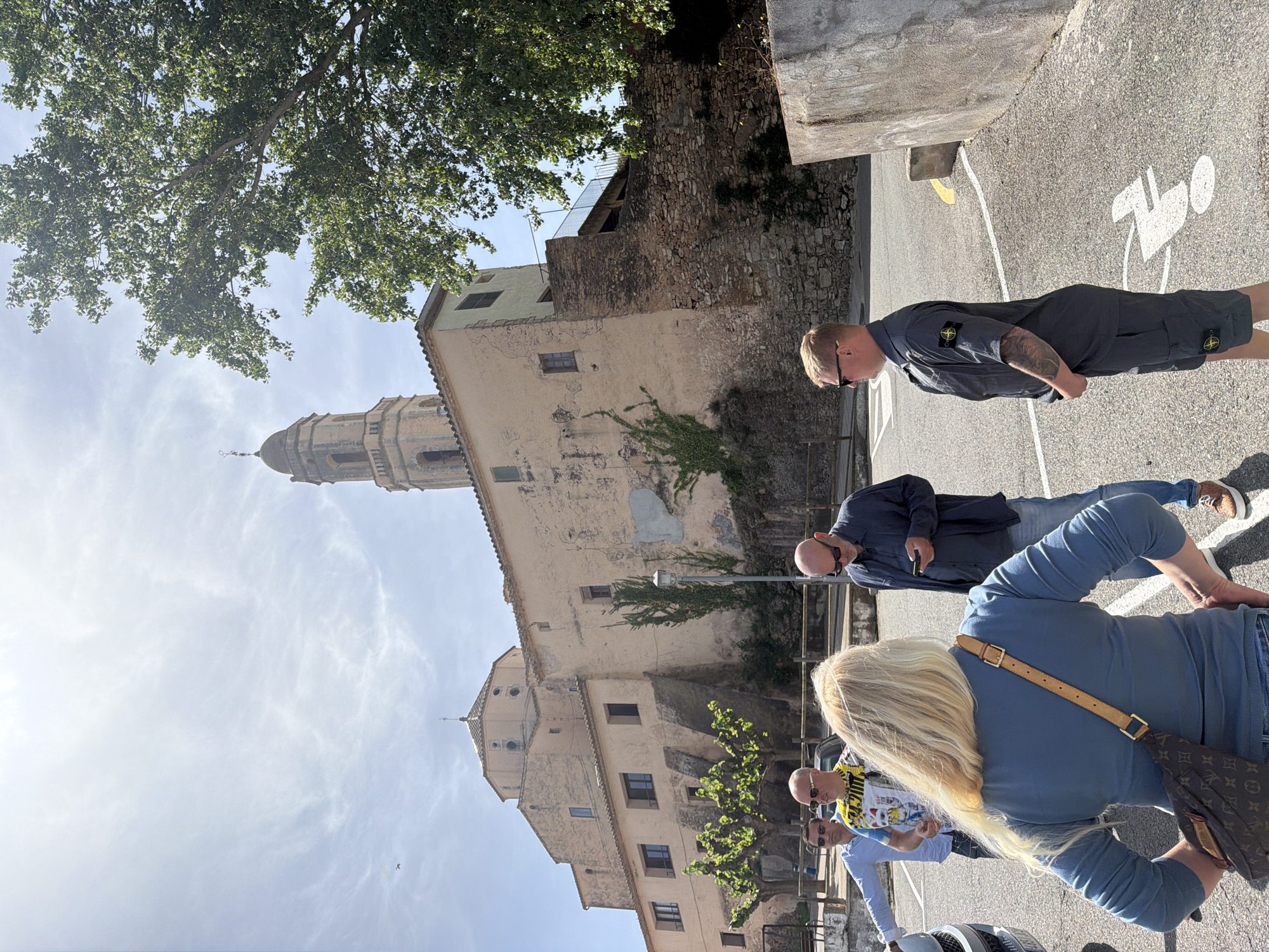 This outdoor scene, set in Bonastre, Spain, features a group of people standing on a paved surface with historical architecture in the background under a bright, sunny sky.
In the background, a tall, light-colored stone church tower with a domed top and a cross at its pinnacle dominates the view, surrounded by other old stone buildings, some with ivy growing on their facades. A large, green-leafed tree is prominent in the upper left of the frame, partially obscuring the sky.
In the foreground and midground, several individuals are casually standing. Closest to the camera, a blonde woman is seen from behind, wearing a light blue long-sleeved top and a brown cross-body bag with a visible Louis Vuitton monogram pattern. To her left, a man in a light blue shirt with a colorful patterned shirt underneath and sunglasses is partially visible. Standing to her right is another man in a dark blue shirt and jeans, wearing sunglasses, seemingly looking downwards. Further back, a man with blonde hair and sunglasses is dressed in a dark grey short-sleeved shirt and matching shorts, both featuring the distinctive Stone Island compass logo patches. He also has visible tattoos on his right arm.
The ground is an asphalt surface marked with white paint, notably a clear disabled parking symbol (a stylized figure in a wheelchair). The sky is light blue with sparse, wispy white clouds, indicating bright daylight and clear weather. The overall impression is a relaxed, casual gathering in a historic setting.