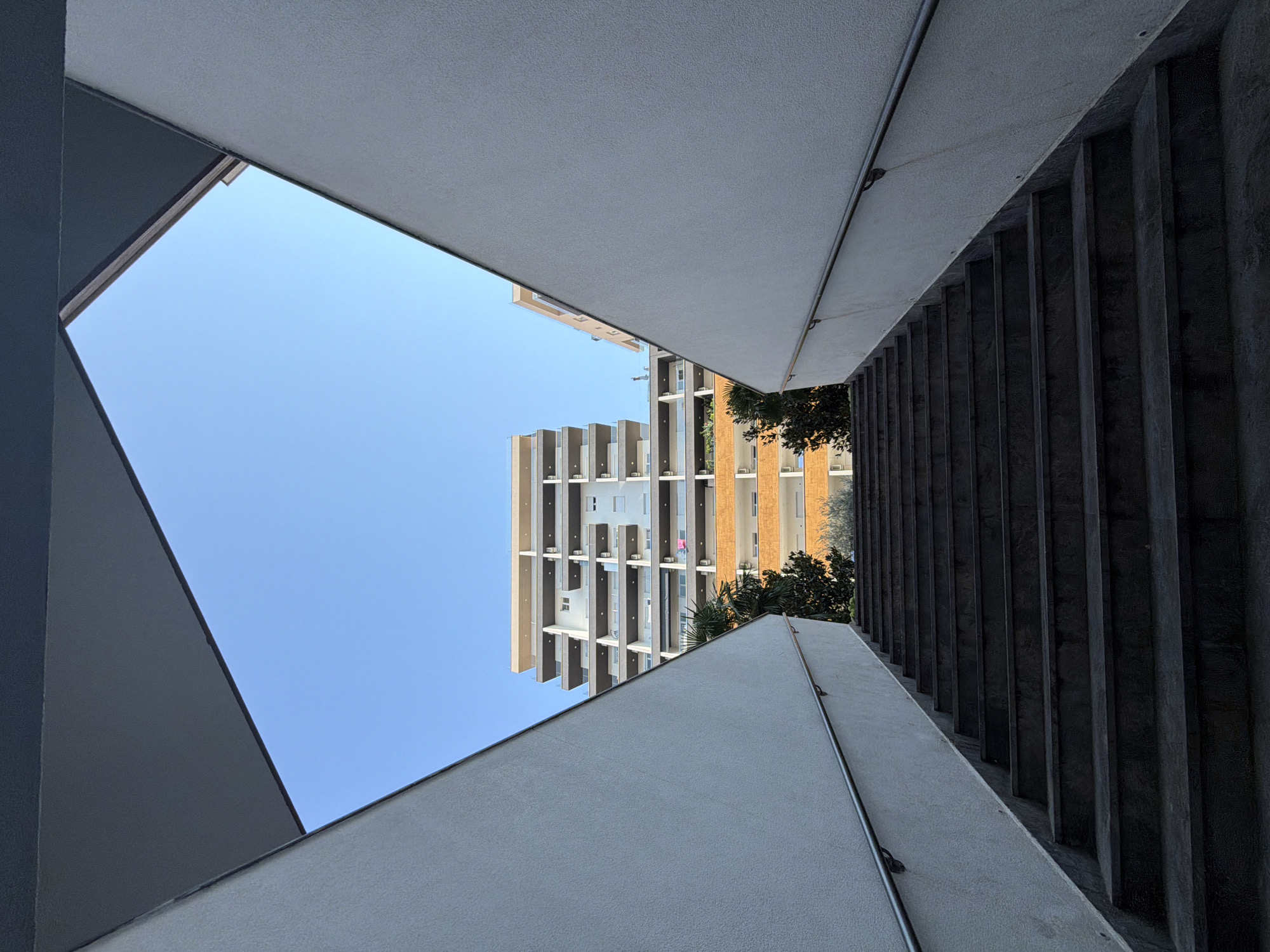 An outdoor, low-angle, full shot shows a concrete staircase leading upwards towards two modern apartment buildings under a clear blue sky. The staircase is framed by two angled, white stucco walls that angle inwards towards the sky, creating a diamond-shaped opening. The staircase steps are dark gray, and a silver metal handrail runs along the left wall. Behind the staircase, the first apartment building is white with multiple balconies and a light-colored top. It appears to be at least 10 stories tall. To the right and slightly behind it is another building, appearing to be a similar height but with orange and white sections and balconies. Green trees and foliage are visible between the staircase and the apartment buildings, including a palm tree to the left. The scene is brightly lit, suggesting it is daytime. The location is likely near Budva, Montenegro, given the architectural style and the presence of palm trees. No people are visible in the image.
