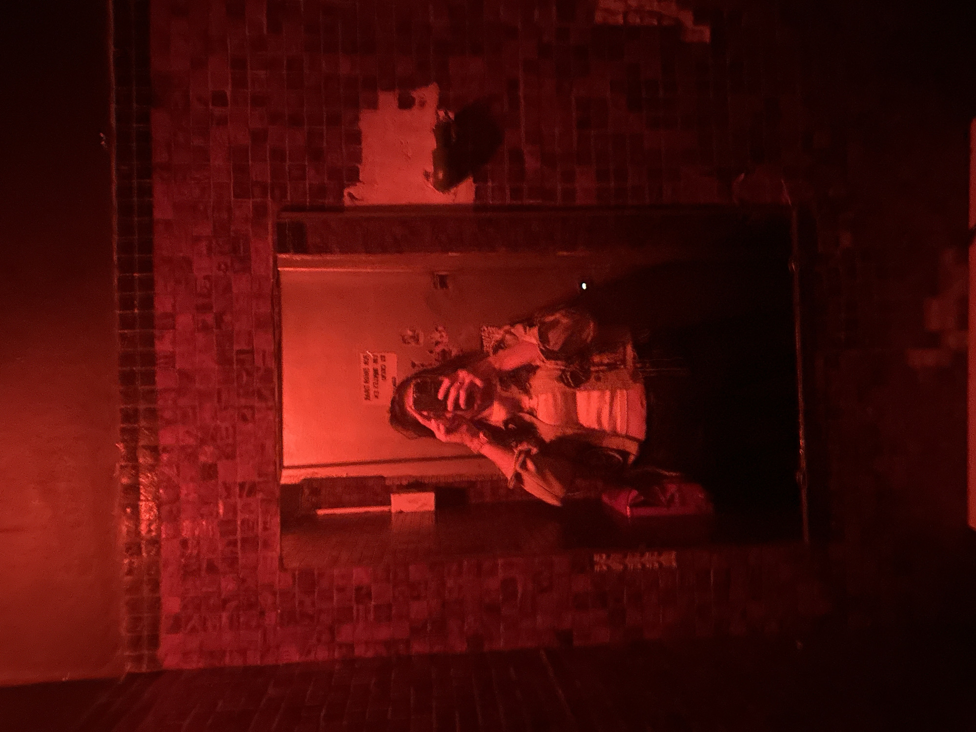 The image captures a vibrant mirror selfie taken in a dimly lit, red-hued indoor space, likely a bar or club in Buenos Aires, Argentina. The main subject is a person, appearing to be a young adult, reflected in a rectangular mirror. They are smiling broadly or laughing, holding a smartphone, presumably to take the photo.
The setting features walls covered in small, square, dark-colored tiles, possibly a mosaic or brick pattern, which are intensely illuminated by a strong red light source, casting a deep crimson glow across the entire scene. Above the mirror, patches of lighter-colored wall are visible, possibly from peeling paint or light reflection. Below the mirror, there's a dark counter or ledge where a red or burgundy bag rests. The red lighting contributes to a lively, somewhat mysterious, and energetic atmosphere, typical of nightlife venues.
On the wall behind the person's reflection, inside the mirror, some white text is visible. Due to the reflection and lighting, it is partially obscured and mirrored, but discernible phrases appear to be in Spanish. The clearest lines read "NO ES CLARO" (meaning "it's not clear") and "MAYOR DE EDAD" (meaning "of legal age" or "adult"). Other words above them are harder to decipher but seem to be part of a set of rules or information. The overall emotion conveyed by the person's expression is one of happiness and enjoyment. The time of day is clearly night, given the artificial, colored lighting.