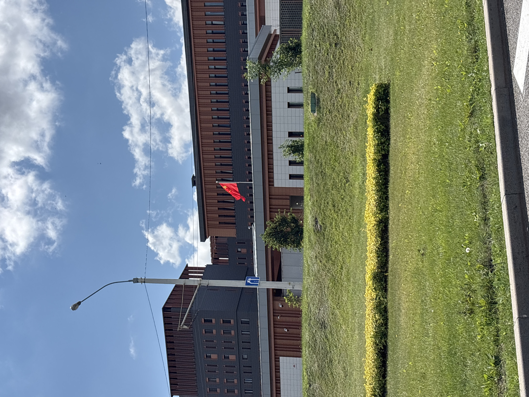 The image captures the exterior of the Embassy of the People's Republic of China in Minsk, Belarus, under a bright, cloudy sky. A light pole with wires stretches from the foreground upwards. The embassy building itself is a modern structure with a dark gray facade, accented by vertical brick-colored elements and numerous narrow windows. A lower section of the building features a white tiled facade with more windows. The Chinese flag, a red banner with yellow stars, flies prominently from a flagpole.
In front of the embassy, a neatly manicured green lawn slopes upwards. A well-defined hedge of yellow-green foliage runs horizontally across the mid-ground of the lawn. The background consists of a bright blue sky populated by scattered white clouds. A traffic sign with a blue background and a white arrow pointing to the right is visible on a pole near the embassy entrance. There are trees and shrubs planted around the building, and security cameras are mounted on the facade. The foreground shows a strip of gray pavement with white markings, suggesting a road or sidewalk. The overall scene depicts a well-maintained and formal institutional setting.