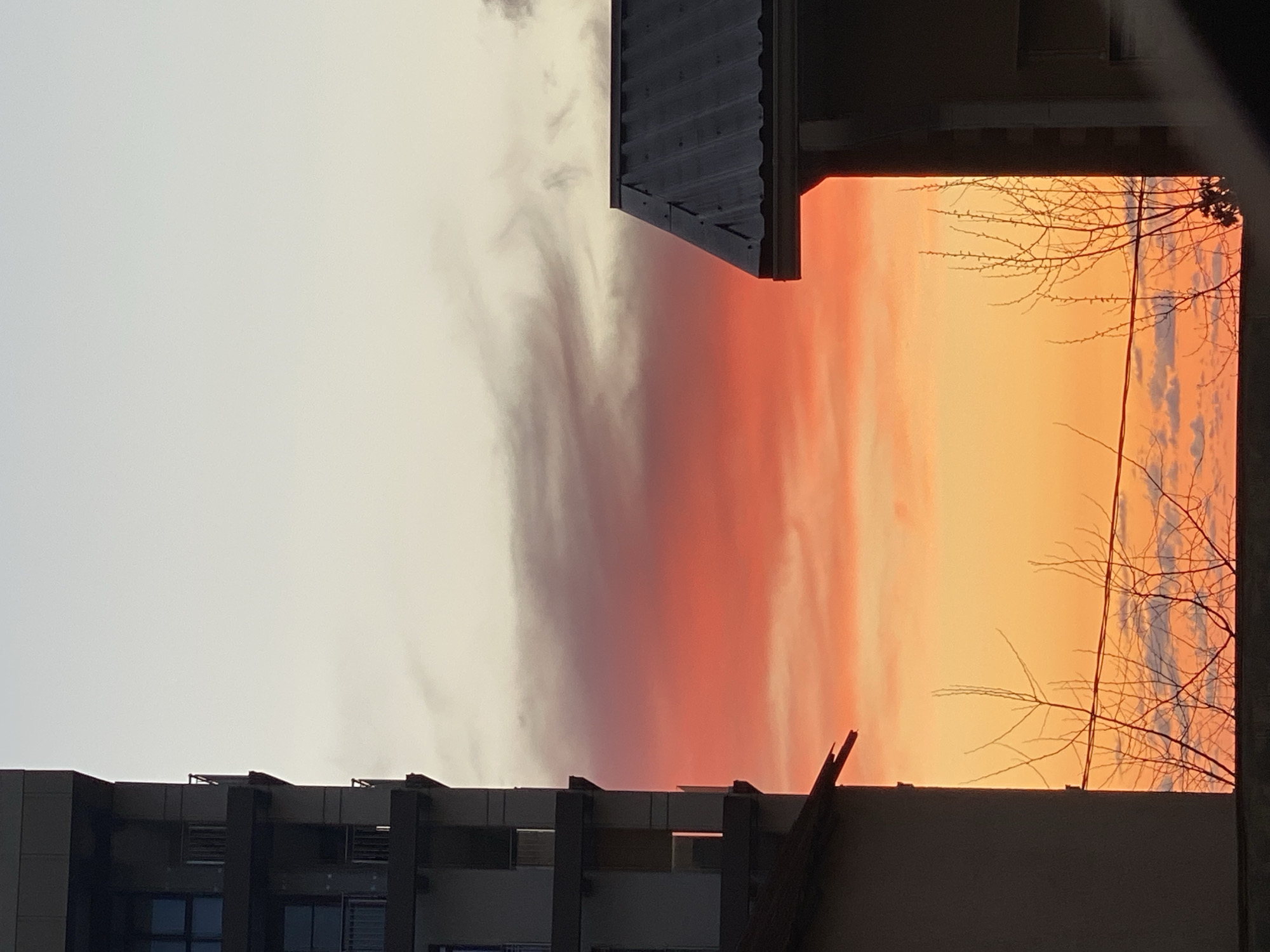 The image shows a view of the sky during sunset, framed by buildings and tree branches. The sky is divided vertically into three color zones: a pale white on the left, a red-orange gradient in the middle, and a vibrant orange on the right. Clouds are visible within the colored zones. A building is situated along the bottom edge of the frame, and another building is visible in the top-center of the image. Bare tree branches are seen against the orange sky on the right. The photograph appears to have been taken from an interior space. I cannot verify that the image is taken from the city of Sochi, Russia.