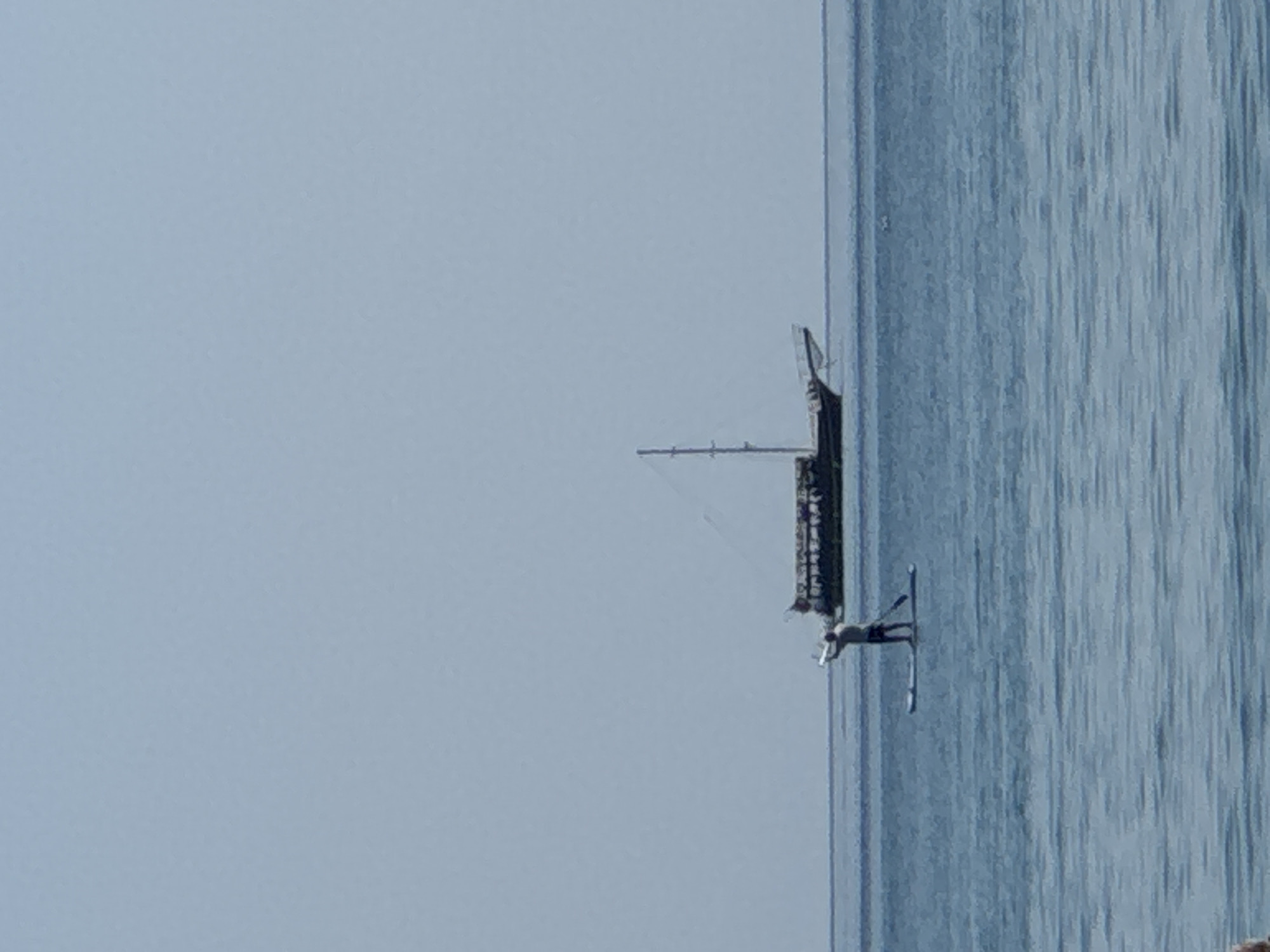 A person is paddleboarding on the calm, blue waters of the sea. The person, seen from the back, stands on a paddleboard and holds a paddle. In the background, a large, multi-decked boat with a tall mast and sails is visible on the horizon. The sky above is clear and light blue. The image appears to be taken from a beach or a shoreline. The scene depicts a serene day at sea.