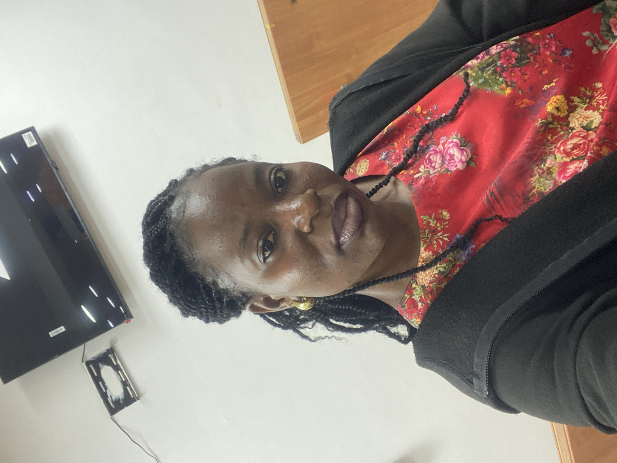 A woman with dark skin and braided black hair is pictured from the chest up. She is wearing a black zip-up jacket over a red floral-print dress. She has on large gold hoop earrings and dark lipstick. Her hair is styled in braids that hang down her front. The background consists of a white wall, a black flat-screen television mounted on the wall, and a wooden surface.