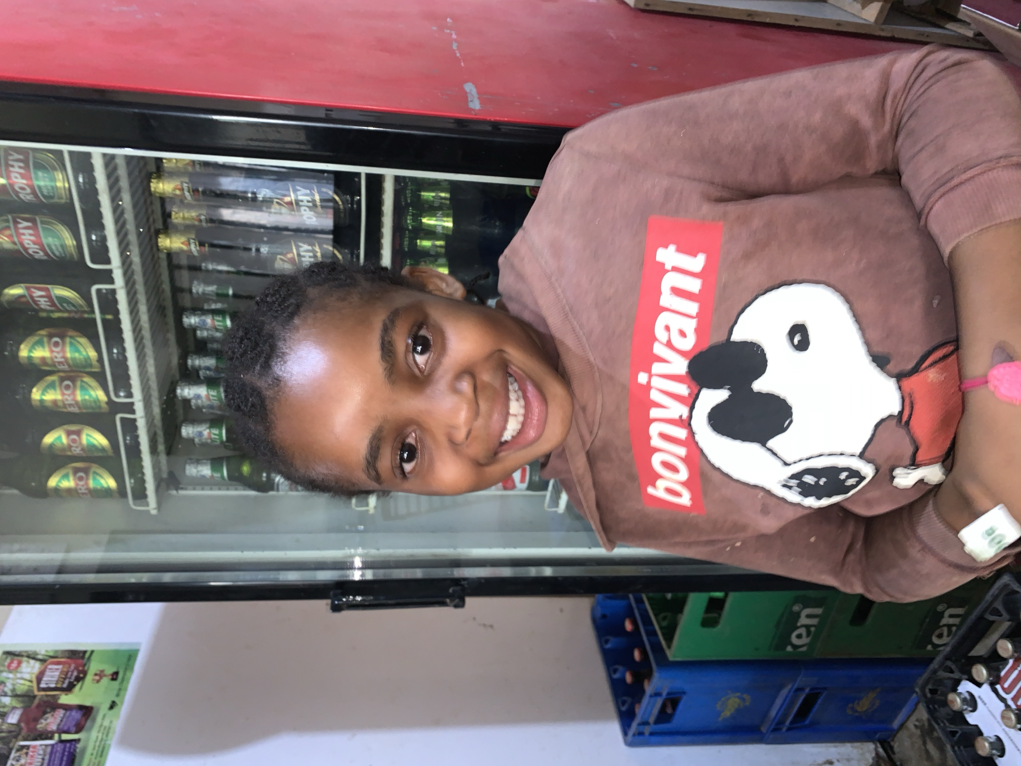 A young Black female with braided hair stands smiling directly at the camera. She wears a long-sleeved, dusty rose or brown sweatshirt featuring a red rectangular label with "bonvivant" in white text, positioned above a cartoon white dog (resembling Snoopy) wearing black sunglasses and a red shirt. On her right wrist, a pink band or object is visible.
Behind her, to the right, is a glass-fronted commercial beverage refrigerator. The top shelf contains several bottles with "ERO" and "TROPHY" visible on their labels. The middle shelf displays more bottles, some with "UTE" and others with "TROPHY" labels. The bottom visible shelf showcases bottles with prominent gold caps and "TROPHY" branding, with additional green-bottled beverages below.
To her left, a white wall is visible, with a poster featuring various products in the upper corner. Below this, stacked blue and green plastic crates are on the floor. Two blue crates are stacked, and next to them, two green crates are stacked, with the partial word "en" (likely "Heineken") visible on their sides. In the bottom left, part of a black crate containing bottles is visible. To the far right of the scene, a red wall or surface shows minor scuffs and marks. The scene is located in the city of New Stadium, Nigeria.