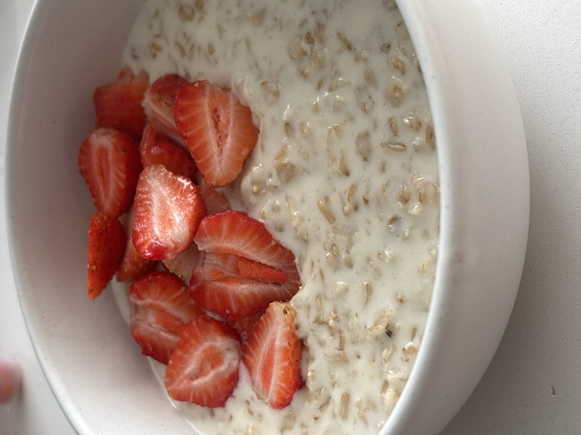 A close-up shot captures a white bowl filled with oatmeal topped with sliced strawberries. The oatmeal appears creamy, with visible oats and a light, milky liquid. Several bright red strawberry slices are arranged on top of the oatmeal, some partially submerged. The bowl itself is a simple white ceramic, seen from a slightly elevated angle, with the right edge of the bowl prominently featured. The background is a plain, light-colored surface, likely a countertop or table. The lighting suggests an indoor setting, possibly during the daytime. No people are visible in the image. The focus is entirely on the food preparation, suggesting a meal, likely breakfast, being presented. The overall impression is one of a simple, healthy, and appetizing dish.