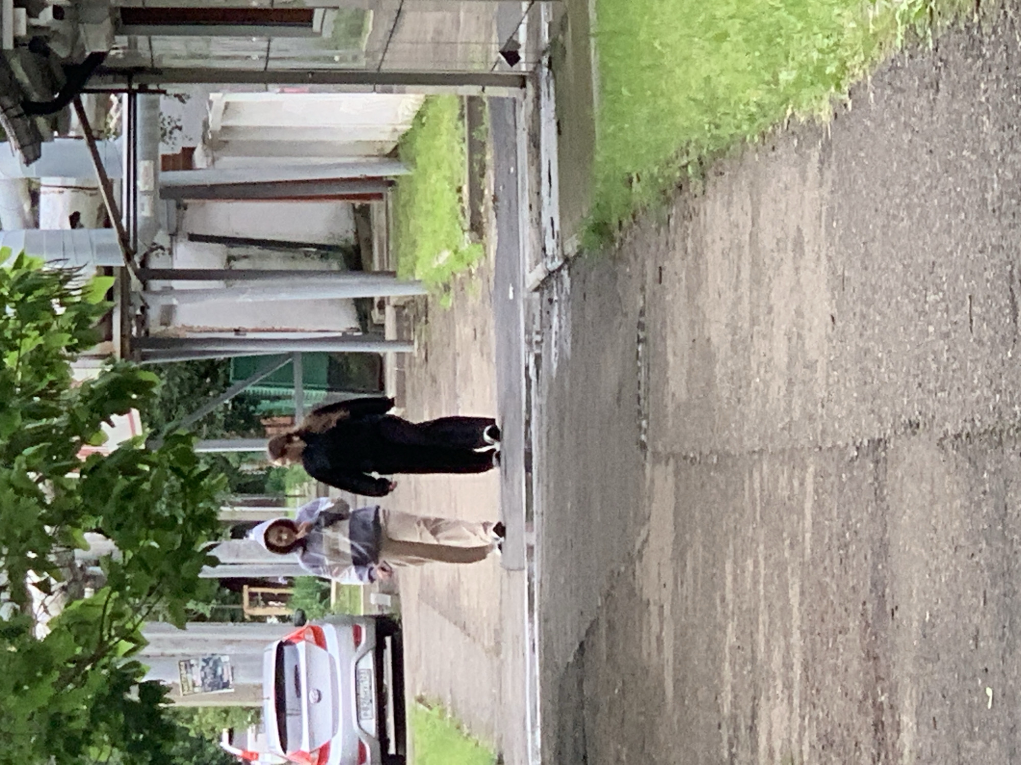 The image captures an outdoor urban scene on a gray asphalt street. Two individuals are walking away from the camera, one in a black outfit and the other in a plaid shirt with beige pants. A silver car is parked near the left edge of the frame. Buildings and pipes run along the left side of the street. Green vegetation and grass border the right side of the path.
