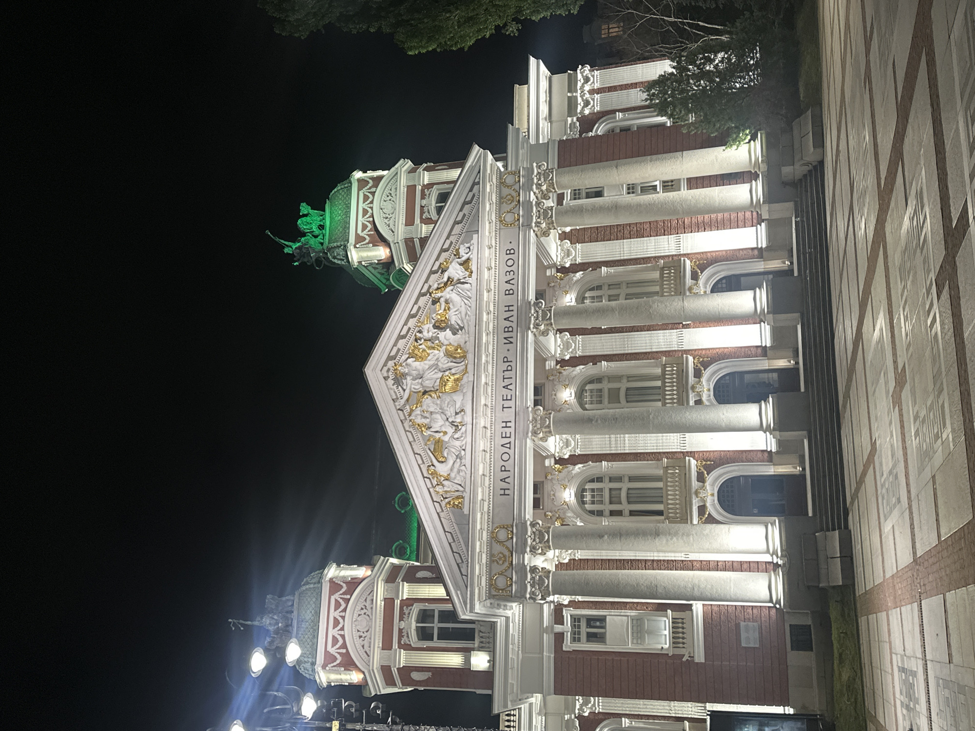 The image shows the Ivan Vazov National Theatre in Sofia, Bulgaria at night. The building is illuminated with bright lights, highlighting its neoclassical architecture. The facade features a central pediment adorned with sculptures and a prominent inscription in Cyrillic. The theater has tall columns and a grand entrance with steps leading up to it. Two towers flank the main structure, topped with domes. One of the domes is lit with green lights and features a statue. The surrounding area is a paved plaza, and a tall tree is visible on the right side of the building. The sky is dark, indicating nighttime. There are no people visible in the image.