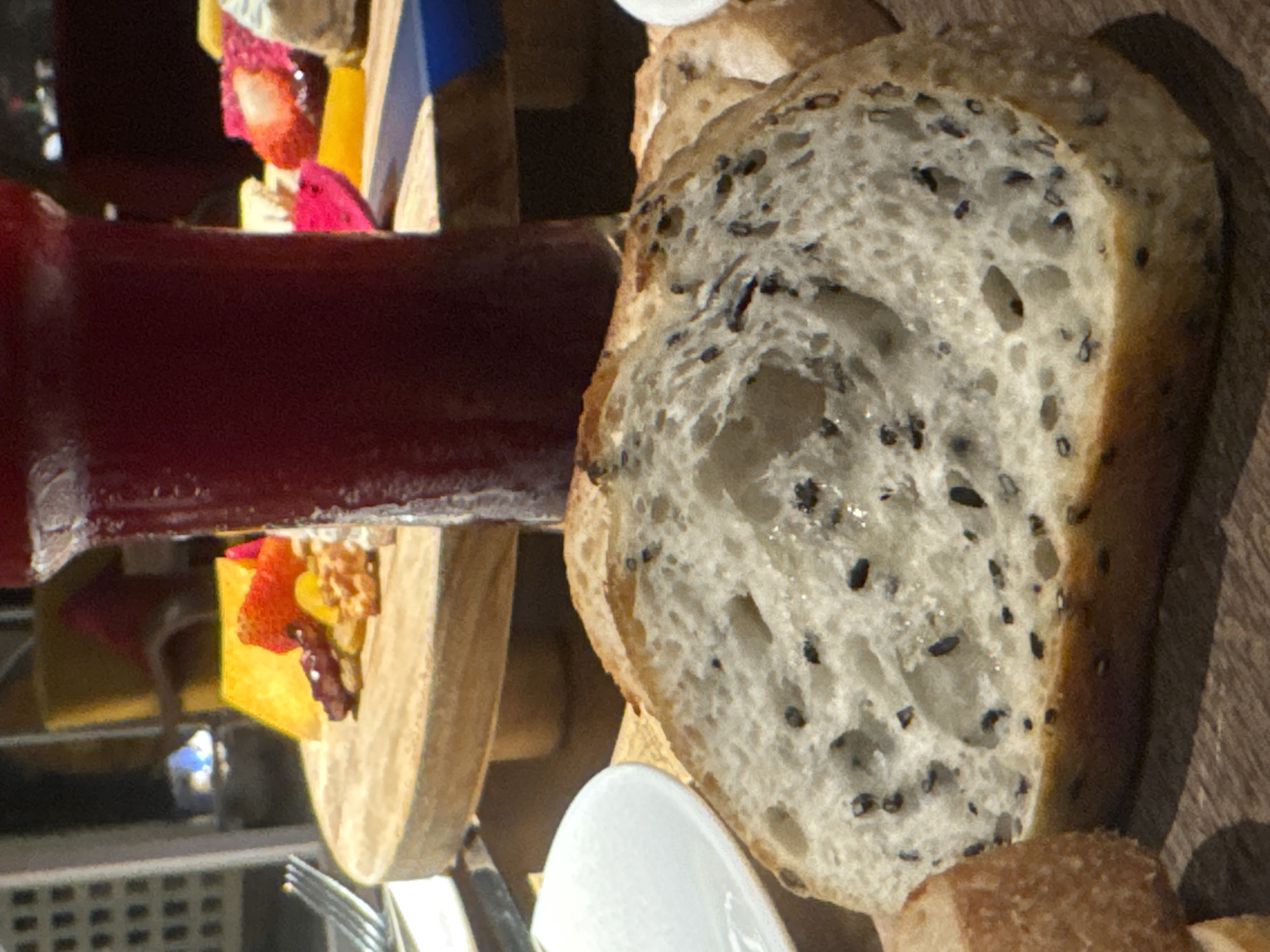The image displays a close-up, side-view of a food presentation, likely a dessert or appetizer platter. The primary subjects are various food items. In the foreground, a slice of rustic bread with black sesame seeds is prominently featured. The bread has a coarse texture and a golden-brown crust. To the left, a dark red liquid, possibly a berry compote or juice, is contained within a clear vessel. Behind the bread and liquid, a wooden board holds several colorful pieces of fruit, including what appears to be sliced strawberries, yellow fruit (possibly mango or pineapple), and possibly some nuts. A pink fruit slice, likely dragon fruit, is also visible. The setting seems to be indoors, with a wooden surface as the backdrop, suggesting a restaurant or home environment. The lighting is somewhat diffused, casting a shadow on the right side of the bread. The image is a still life composition focused on the visual appeal of the food. There is no discernible text in the image. The context provided, Hà Nội, Vietnam, suggests this food presentation is part of Vietnamese cuisine or dining culture.