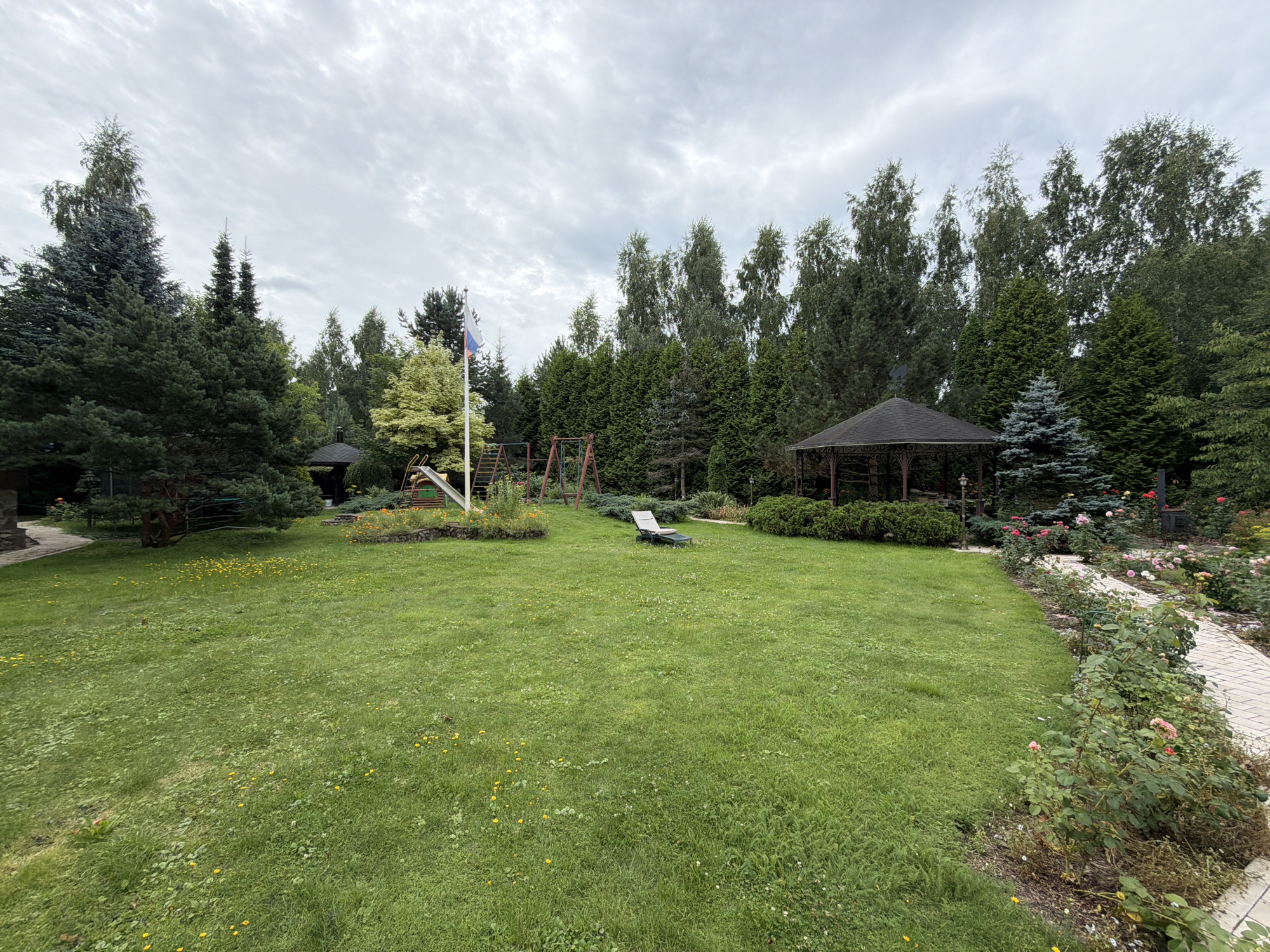 The image shows a cloudy day in a backyard with a lush green lawn. In the background, a row of tall trees lines the property. To the left, there is a children's playground with a slide and swings. A gazebo is visible in the mid-ground, and a rose garden with a paved walkway is on the right. A single lounge chair is placed on the lawn. The location is identified as Sannikovo, Russia.
