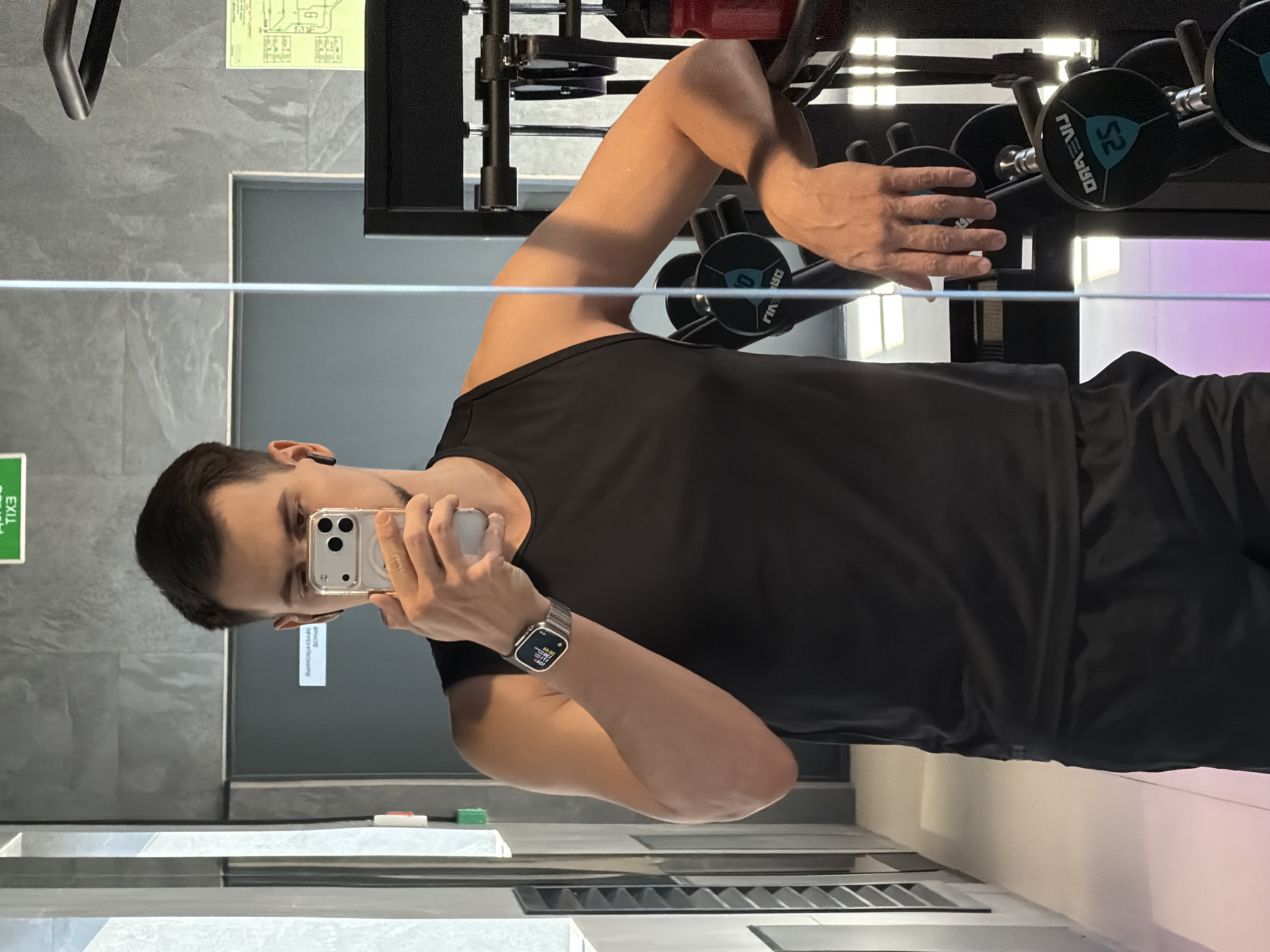 The image, taken in a gym setting in Krasnoyarsk, Russia, shows a man taking a mirror selfie. He is positioned in front of a mirror, looking directly into his smartphone, which he holds up with his right hand. The man is wearing a black sleeveless athletic top and black shorts. On his left wrist, he has a silver-banded smartwatch with a visible digital display. A black earbud is visible in his left ear.
Behind the man, visible in the mirror's reflection, is gym equipment, specifically a rack holding multiple black dumbbells with blue markings. One dumbbell clearly shows the number "52" and partially legible text that appears to be "GRAEVU" (likely mirrored). The background features grey textured walls. A white horizontal bar or shelf runs across the middle of the frame, possibly part of the gym's interior design or a shelf in front of the mirror.
On the left side of the reflection, a green "EXIT" sign (mirrored as "TIXE") is visible, along with a yellow diagram or map on the wall above it, though its details are unreadable. The overall lighting is bright and artificial, typical of an indoor gym. The activity is a casual mirror selfie, with no strong emotional cues displayed by the man beyond a neutral expression focused on his phone. The time of day cannot be determined as the setting is indoors.