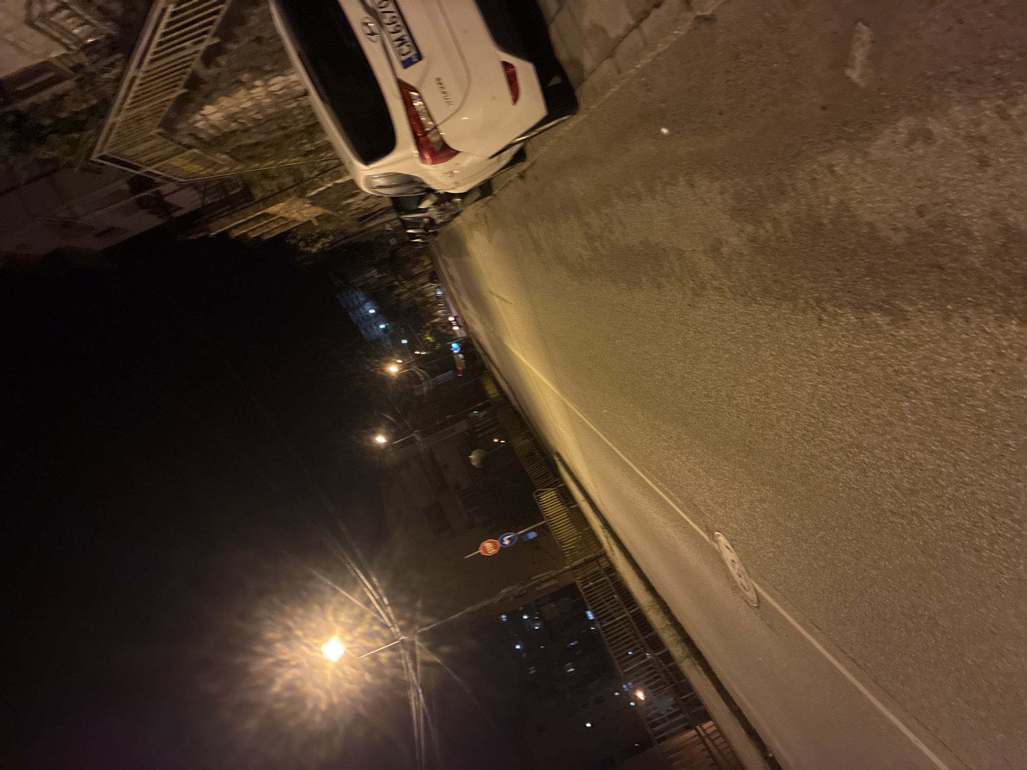 The image depicts a nighttime street scene in Vlahovo, Smolyan Province, Bulgaria, illuminated by artificial streetlights. The perspective is low-angle, looking down a two-lane asphalt road that gently curves to the left in the distance. The road surface, which occupies the majority of the frame, is a dark grey asphalt with a warm, yellowish glow from the streetlights. A single white solid line marks the right edge of the visible road lane. A circular manhole cover is visible on the road surface in the mid-ground.
On the right side of the road, a concrete sidewalk is present, where a white Hyundai passenger car (license plate `CM 6676`) is parked, facing away from the viewer. The "CM" on the license plate identifies its registration origin as the Smolyan region of Bulgaria. Behind the parked car, a stone wall and multi-story buildings with balconies and windows are visible, along with metal railings and stairs suggesting an elevated or terraced structure.
On the left side of the road, a bright streetlight with a pronounced starburst flare illuminates the foreground and mid-ground. Further down this side, a red octagonal STOP sign is mounted on a pole, with a blue circular mandatory direction sign below it. Metal railings, possibly indicating a bridge or elevated walkway, are also present on the left. In the background, additional streetlights and dimly lit buildings contribute to the urban environment. No people are visible in the scene.