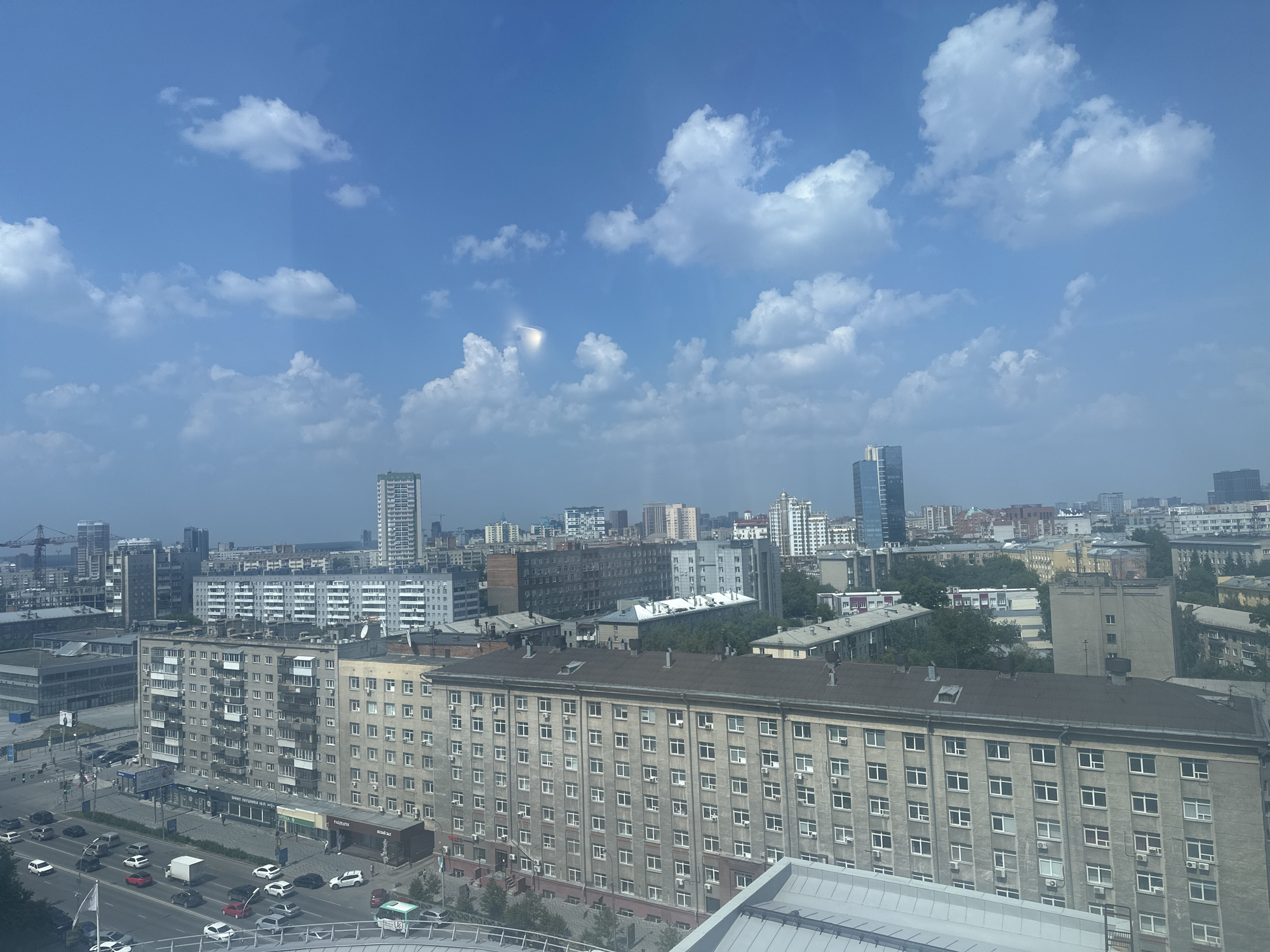 This is a high-angle, wide shot of a city with a clear blue sky and scattered white clouds. The foreground features a street with numerous cars of various colors, including white, black, red, and silver. A white truck is visible on the street. There are several buildings in the midground, most of which are multi-story apartment buildings with many windows. Some of these buildings have balconies. In the background, there are more buildings of varying heights, including some modern high-rises and older Soviet-era apartment blocks. Trees are visible in some areas between the buildings. The overall impression is of an urban environment on a sunny day.
The location of this image has been identified as Novosibirsk, Russia. Specifically, the cluster of older, block-style apartment buildings with a large, uniform facade facing the street is characteristic of the architecture found in many residential areas of Novosibirsk built during the Soviet era. The presence of both older and newer high-rise buildings suggests a city that has undergone development and modernization. The street scene with traffic and pedestrian activity further reinforces this identification as a typical urban setting within Novosibirsk.