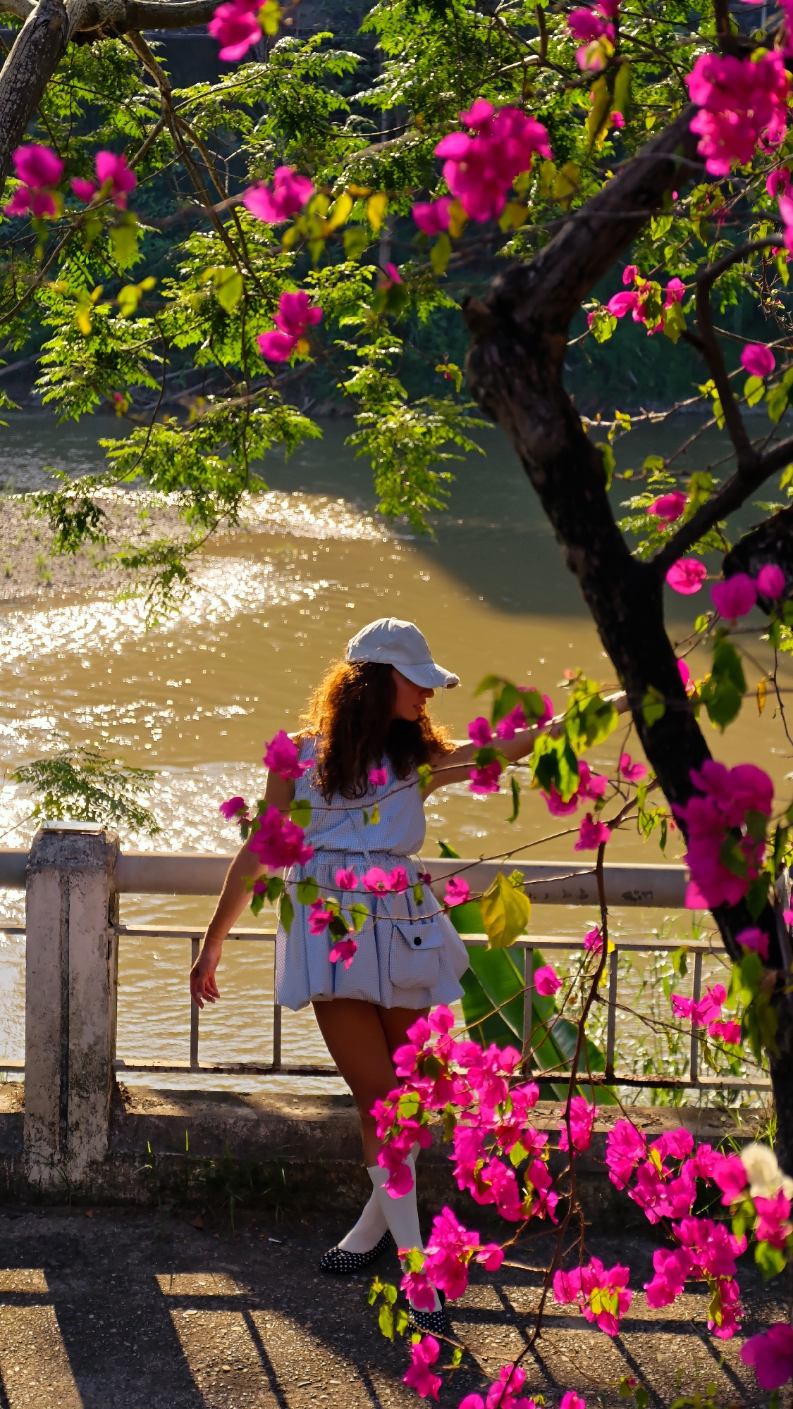 A young woman stands on a stone patio near a railing overlooking a river. She is wearing a light blue dress, white socks, and black and white polka-dotted shoes. Her brown hair is visible under a light-colored baseball cap. She is facing the river and touching the railing with her right hand. Pink bougainvillea flowers frame the scene. The river reflects sunlight, and trees are visible on the opposite bank. The scene is in Lao Cai Ward, Vietnam.