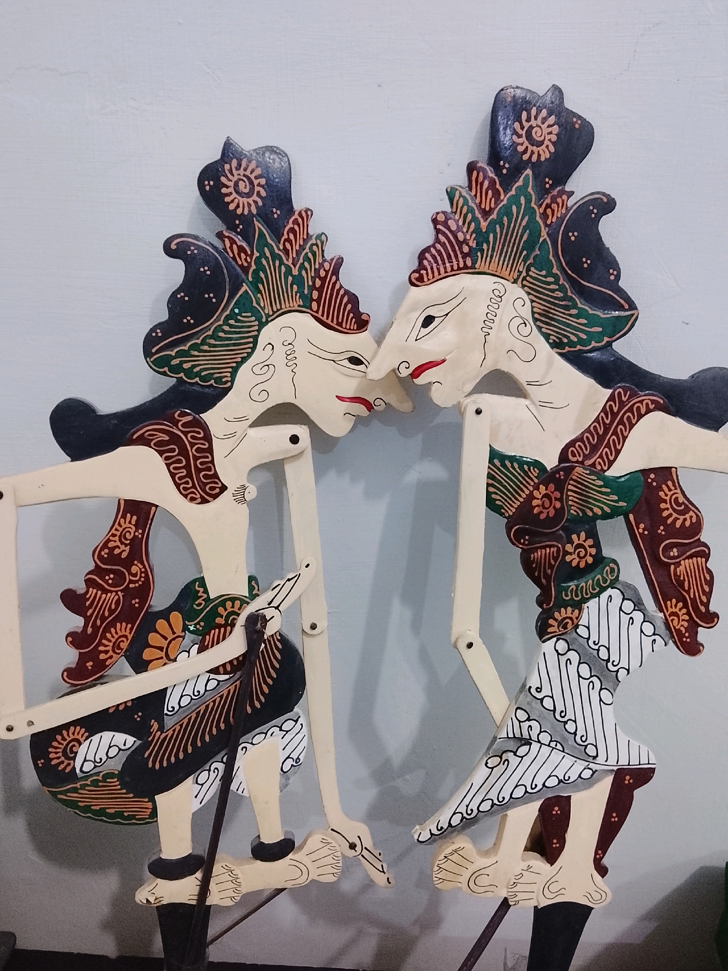 Two traditional Javanese wayang golek puppets, carved from wood and painted with vibrant colors, are positioned face-to-face with their lips almost touching, against a plain, light-colored wall. Each puppet features elaborate headdresses in shades of black, green, red, and yellow, with intricate patterns. Their faces are pale with defined eyes and red lips. They wear ornate garments depicting various motifs, including floral designs and batik-like patterns in brown, black, green, and white. The puppets are articulated with wooden rods and joints, suggesting they are ready for performance. The setting appears to be indoors, possibly a home or a cultural exhibition space in Slawi, Tegal Regency, Central Java, Indonesia, given the style of the puppets and the commonality of such cultural artifacts in the region.