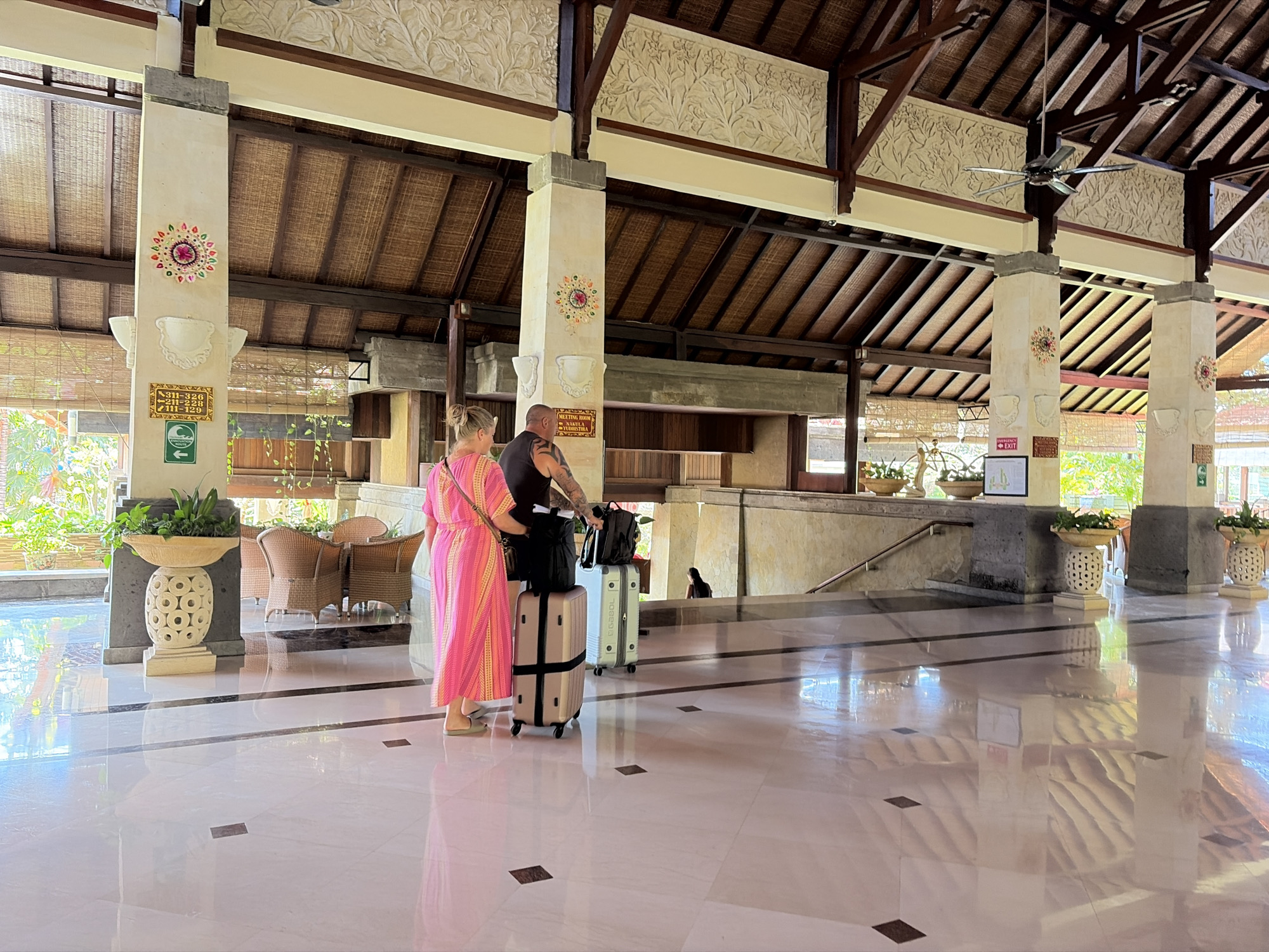 In the lobby of the Grand Hyatt Bali in Kuta Selatan, Indonesia, a woman in a pink dress and a man in a black tank top stand with their luggage. The woman, seen from the back, is wearing light-colored sandals. The man, also facing away, has tattoos on his arms. They are positioned on the polished marble floor, which reflects the opulent architecture of the lobby. In front of them are two suitcases: a light-colored, upright model and a silver, larger one. Behind them, a staircase ascends, and in the background, the lobby features high, thatched ceilings with intricate carvings, large pillars adorned with colorful mandala-like designs, and woven blinds. Various potted plants and rattan furniture are visible, contributing to the tropical ambiance. A ceiling fan is mounted on the wooden beam structure. The scene appears to be a moment of arrival or departure, with the couple ready to move through the resort.