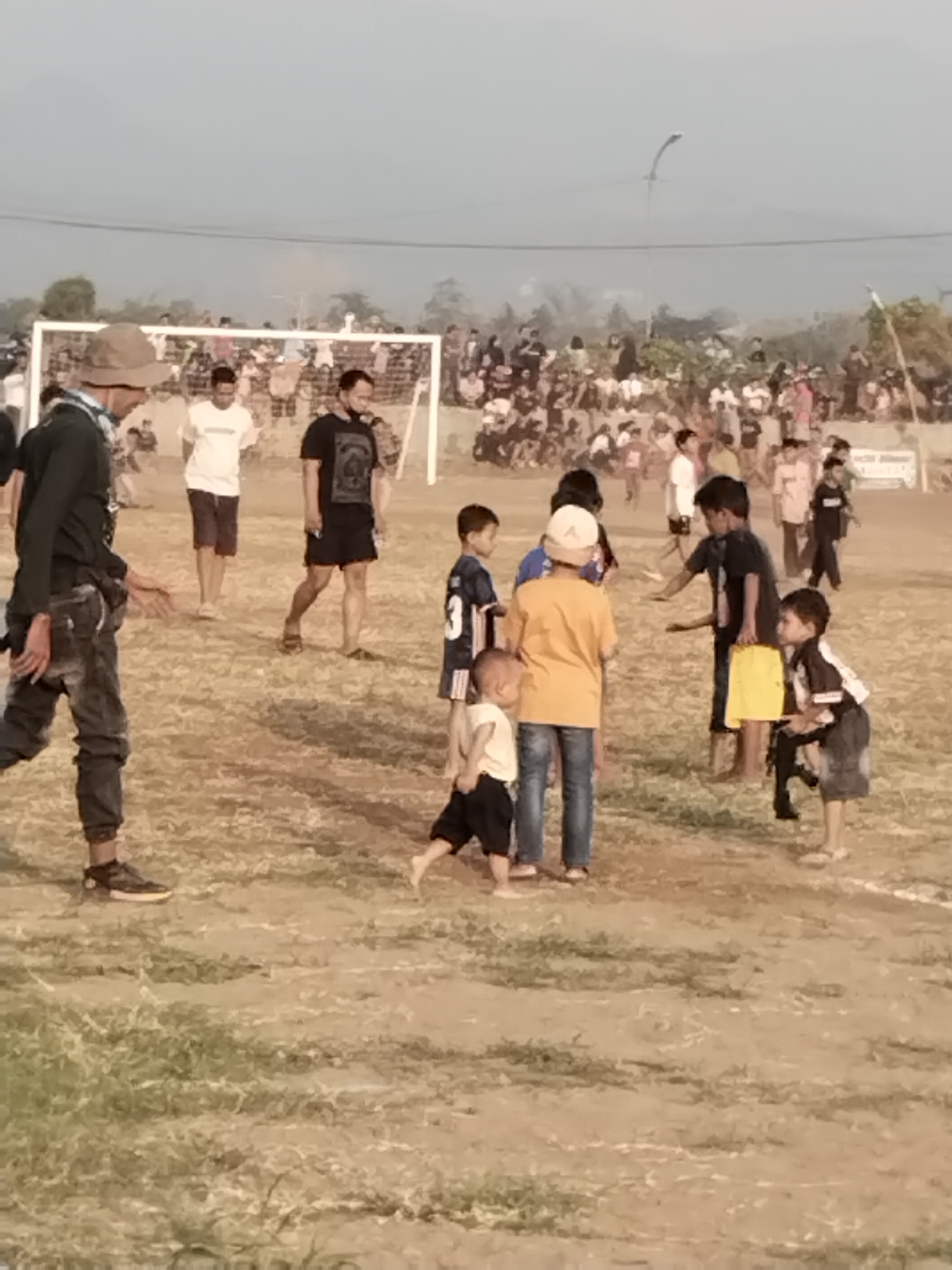 This image depicts an outdoor scene, likely a soccer field, in Margasari, Indonesia. A crowd of spectators is visible in the background, some standing and some seated, behind a white soccer goal. In the foreground, several individuals, primarily children and young adults, are present on the field. One young boy is wearing a white t-shirt and black shorts. Another boy, wearing a yellow t-shirt and blue jeans, stands with his back to the camera. A boy in a black and white soccer jersey with the number '3' is also visible. An adult male in a dark long-sleeved shirt and jeans is walking from left to right. Another adult male in a black t-shirt and shorts stands near the goal. The ground appears to be dirt or dry grass. The sky is overcast, and a light pole with wires is visible in the upper portion of the image.