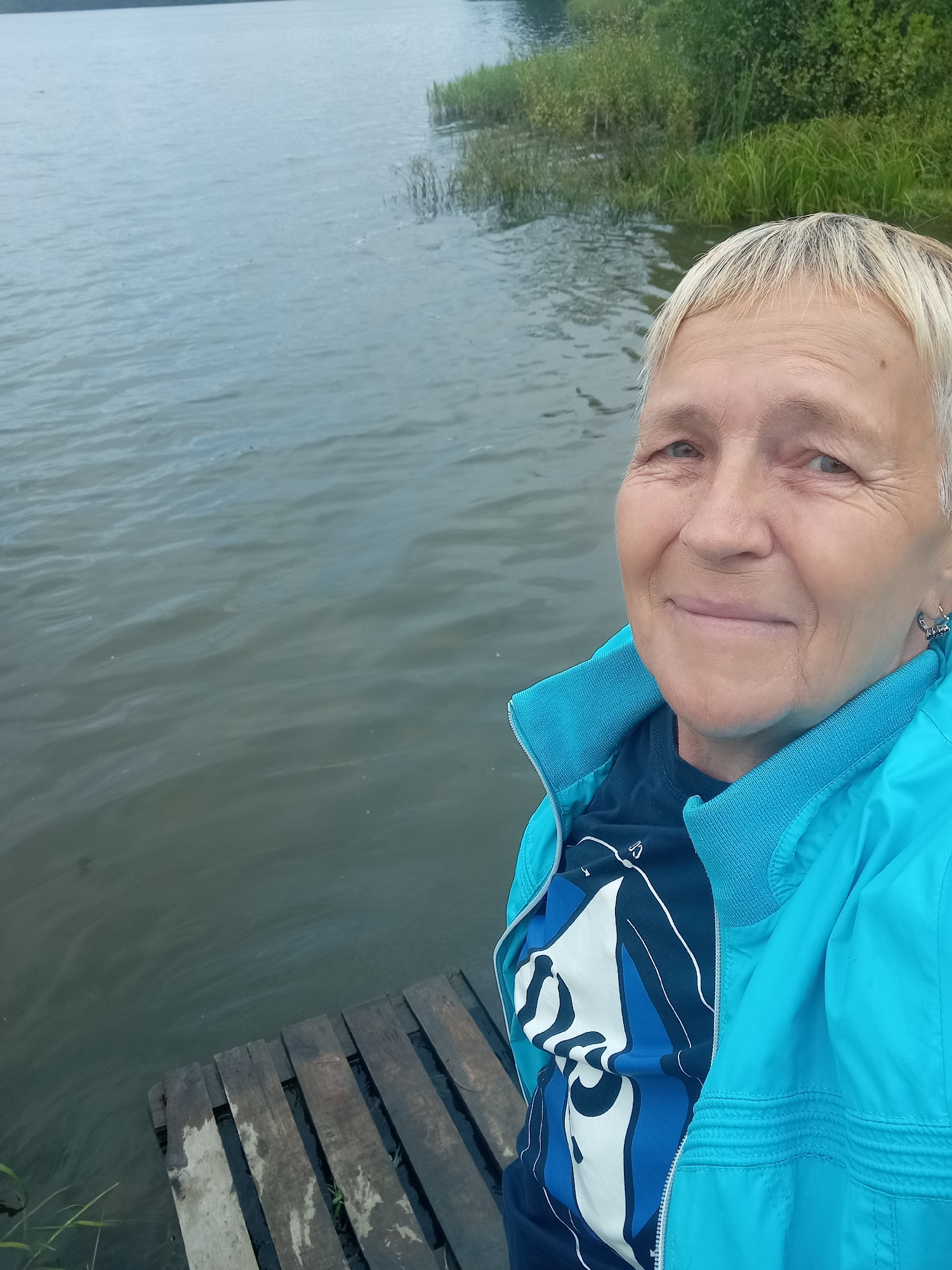 A woman with short blonde hair is standing on a wooden pier by a body of water, with greenery on the far bank. She is wearing a blue jacket over a dark blue t-shirt with a white and blue graphic design. She is smiling at the camera. The image appears to have been taken outdoors in a natural setting.
