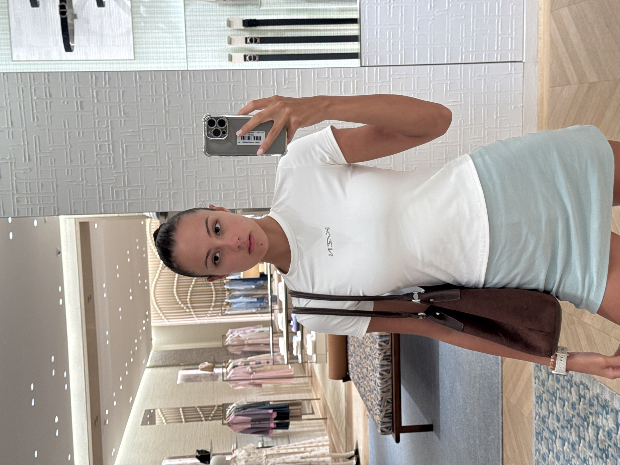 A woman takes a selfie in a retail store with her smartphone. She is wearing a white t-shirt and a light blue skirt. She is holding a brown purse in her left arm. The background shows clothing racks and displays. There is a patterned bench in the middle ground. The floor is tiled. The woman is standing on a blue and white patterned rug. The store appears to be well-lit with overhead lighting. The location is identified as Wichit, Thailand.
