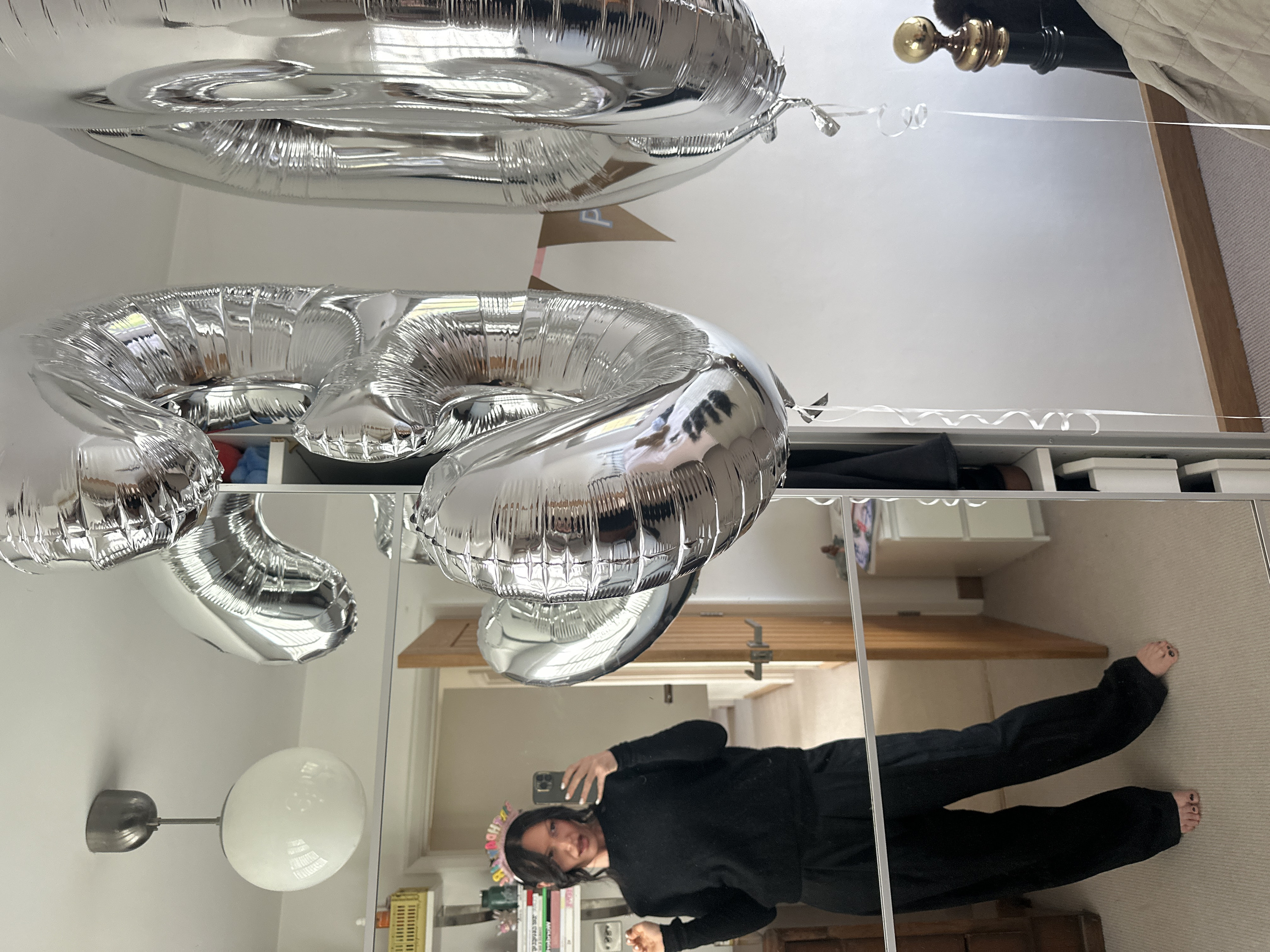 An indoor scene within a residential property in Tunbridge Wells, United Kingdom, is depicted. The primary subject is a female individual reflected in a large, floor-length mirror, taking a selfie with a smartphone held in her right hand. She wears a dark long-sleeved top and dark wide-leg trousers. A "HAPPY BIRTHDAY" headband adorns her head. Her bare feet, with dark nail polish, are visible on a light-colored carpet.
In the foreground, two large silver foil balloons shaped as the numbers "5" and "0" are prominently displayed. The "5" balloon is positioned to the left, partially overlapping the "0" balloon to its right. Both balloons have white curling ribbons extending downwards. Behind the balloons, a section of a brown banner with the letter "P" from a "HAPPY BIRTHDAY" garland is visible.
The mirror reflects a section of the room: to the left, a white shelving unit holds books and various objects, including a yellow crate and a power outlet. A light-colored wooden door frame and a slightly ajar wooden door lead to an adjacent room or hallway. A ceiling light fixture with a spherical white glass shade is mounted on the white ceiling in the upper-left. To the far right, a dark bedpost with a brass finial is partially visible, alongside a beige duvet or blanket. The room features white walls and a light-colored wooden baseboard running along the floor. The vertical frame of the mirror/wardrobe unit reveals internal shelving compartments containing various items.