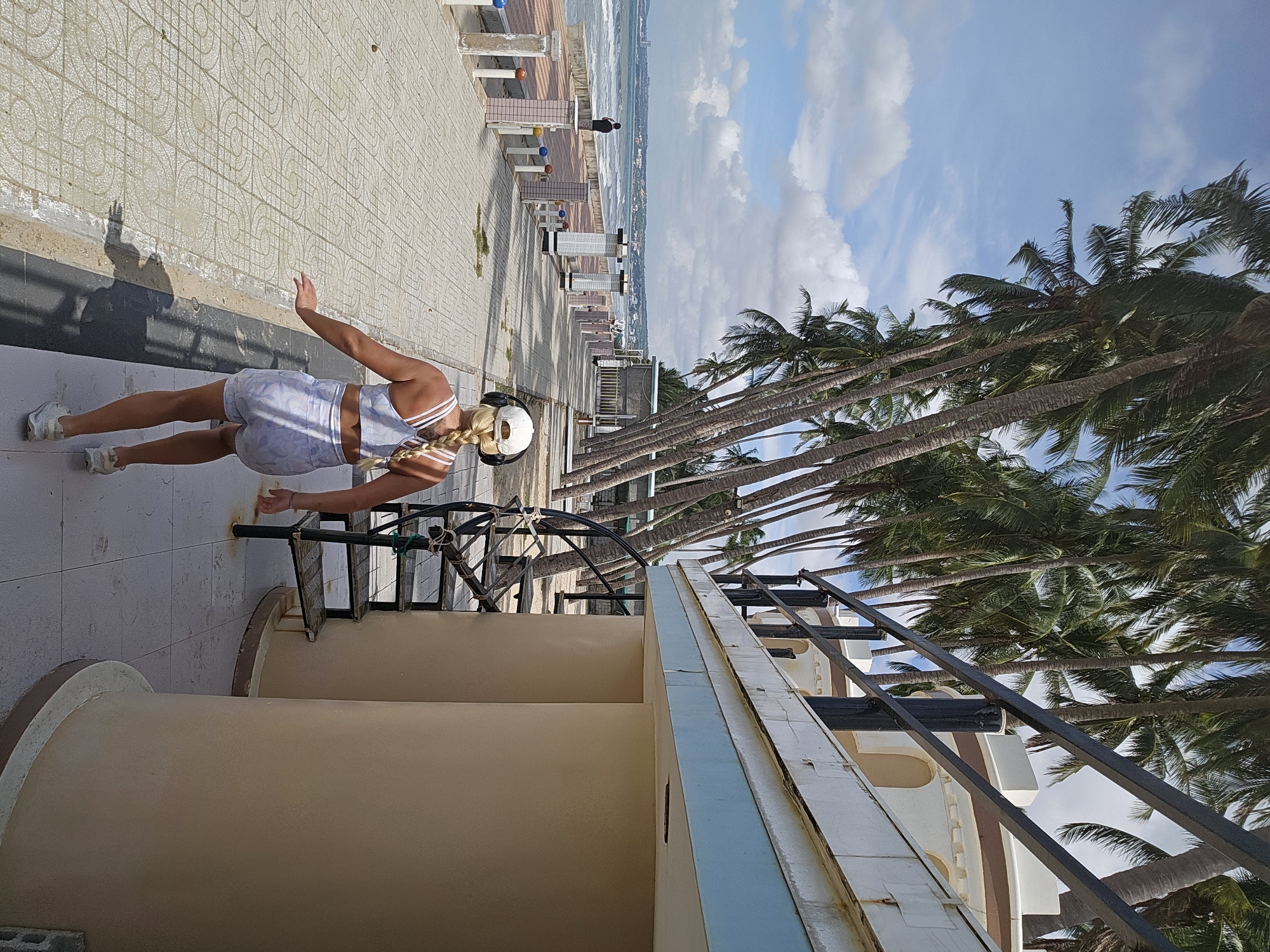 A woman in athletic wear, including a light-colored patterned sports bra and shorts, performs a plank exercise outdoors. She wears white sneakers and headphones. Her blonde hair is braided. She is positioned on a tiled walkway next to a beige structure with a black railing and stairs. In the background, a palm-lined promenade overlooks a beach and the ocean. The sky is blue with scattered white clouds, suggesting daytime with pleasant weather. The scene appears to be a tropical resort or public space, possibly a beachfront area in Mũi Né, Vietnam, given the prominent presence of palm trees and the architectural style. The woman's posture indicates physical activity and focus.