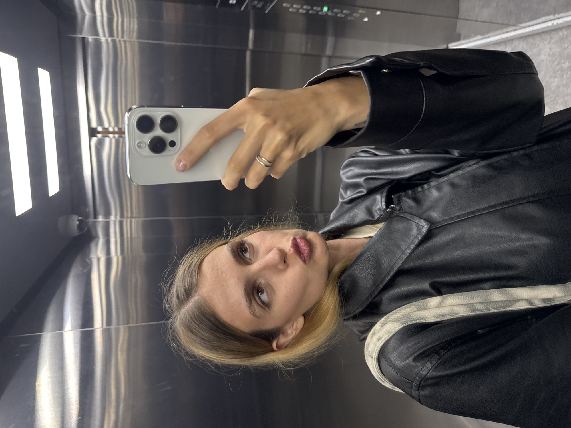 A woman is taking a selfie with a white smartphone with three rear cameras inside an elevator. The elevator is silver with fluorescent lighting, and the woman is wearing a black leather jacket with a white stripe over the shoulder. She has a ring on her left hand.