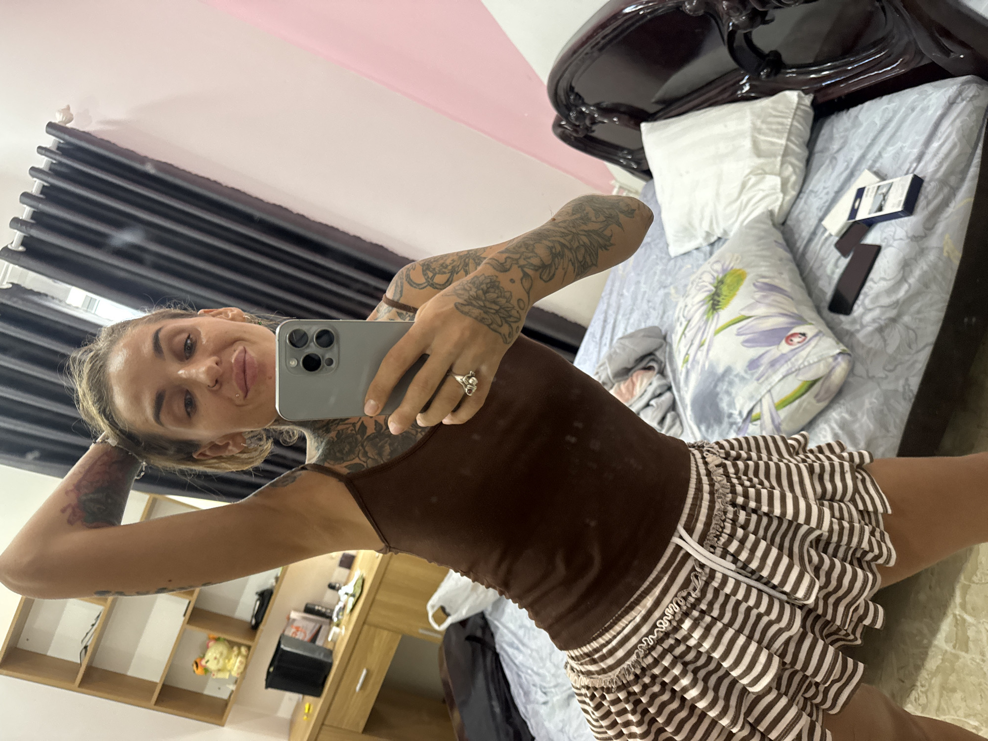 A young Caucasian woman with blonde hair, styled in a bun, is taking a mirror selfie in what appears to be a bedroom. She is wearing a brown tank top and a brown and white striped skirt. She has numerous tattoos on her arms and chest. Her left arm is raised, with her hand behind her head. She is holding a silver smartphone in her right hand. In the background, a wooden shelf unit with various items is visible, along with a black curtain and a pink wall. The room also contains a bed with pillows and what appears to be a wooden bed frame. The overall setting suggests a casual, personal space within a home.