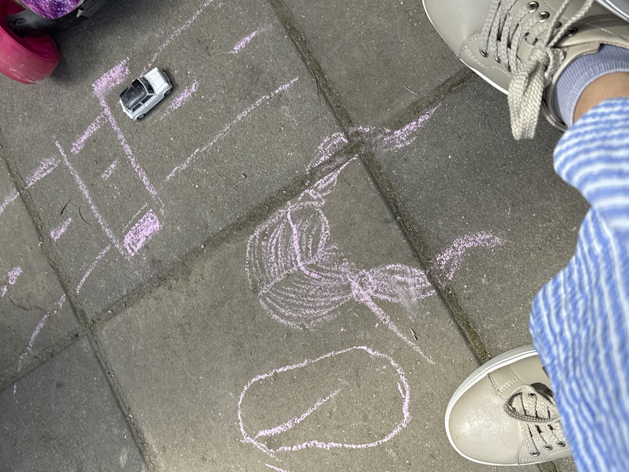 The image shows a sidewalk in Unknown, Unknown, with children's chalk drawings in light purple. A hopscotch grid, a flower, and an oval shape are visible. A toy car sits on the hopscotch grid. A person wearing light-colored sneakers and blue and white striped pants stands on the sidewalk. A child's roller skate is partially visible.