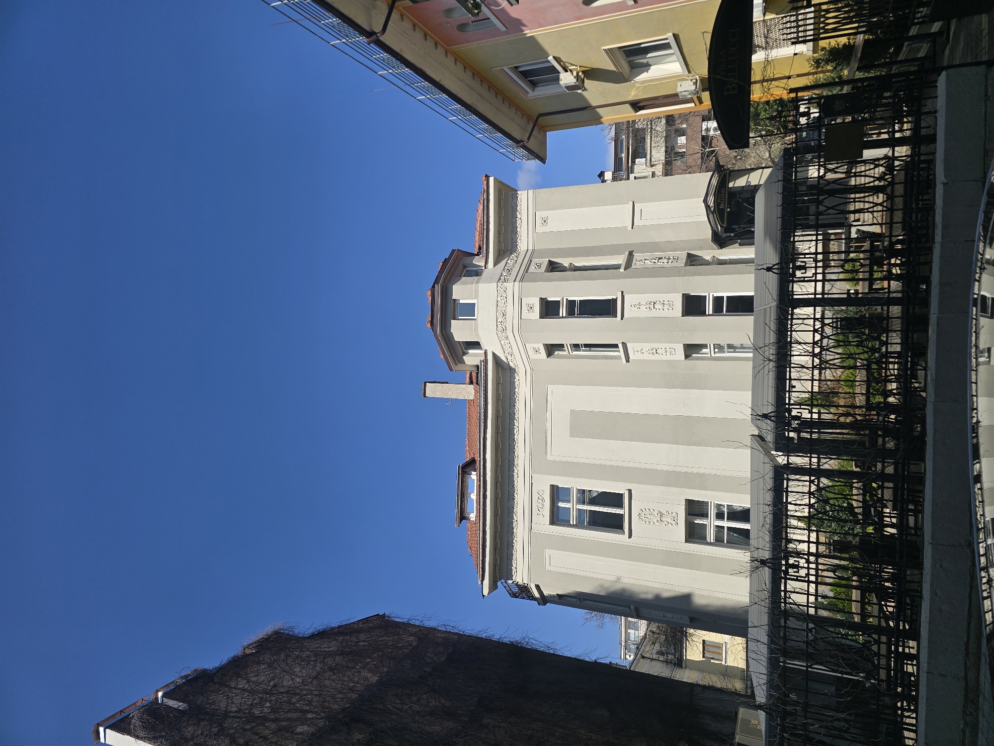 The image displays the exterior of the "Villa Bellucci" building, located at ul. "Mali Bogdan" 1, 1504 Sofia Center, Sofia, Bulgaria. The central three-story structure presents a light gray facade with ornate decorative moldings beneath its rectangular, white-framed windows. It features a terracotta-tiled roof with a prominent octagonal turret and a single dormer window, alongside a visible chimney stack.
To the immediate left, a dark building wall is extensively covered with a dense network of bare vines. To the right, a multi-colored building, primarily yellow and pink, exhibits a modern metal railing along its roofline and two visible external air conditioning units.
In the foreground, a dark wrought-iron fence with intricate scrollwork runs horizontally, separating the viewer from an outdoor dining area. This area is furnished with multiple dark tables and chairs, protected by gray awnings or canopies, and incorporates various green plantings. A partial reflection of the fence and foreground elements is visible at the very bottom edge of the image. The scene is illuminated by direct sunlight under a clear, unobstructed bright blue sky. No individuals are visible.