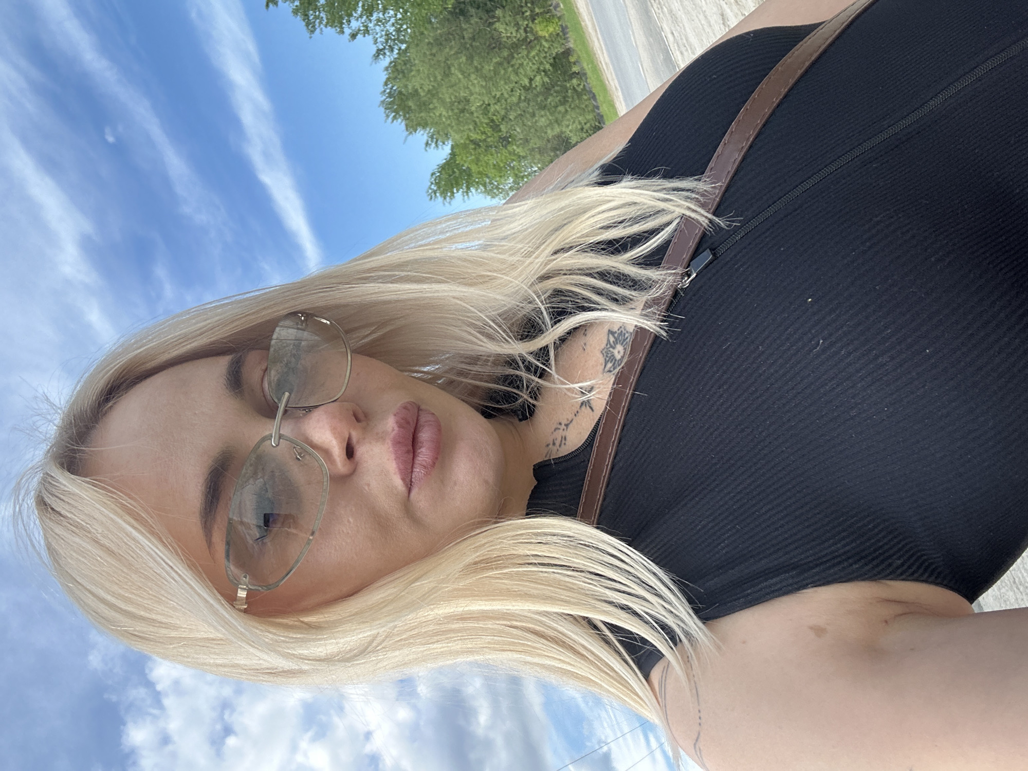 The image captures a close-up, outdoor selfie of a blonde woman. She is positioned centrally, looking directly into the camera with her lips slightly pursed. She wears rimless, lightly tinted sunglasses that reflect the sky and faint structural elements. Her hair is shoulder-length and straight. Tattoos are visible on her neck/upper chest (appearing as small script or symbols), her right collarbone area (a circular floral design), and faint lines on her left upper arm. She wears a black, ribbed, sleeveless top featuring a central zipper. A brown leather strap, possibly from a bag, crosses her chest from her right shoulder. The background features a bright blue sky with scattered white clouds, and green trees are visible to the right. A light-colored ground surface, resembling a road or path, is present in the lower right background. The scene is located in Unknown, Unknown.