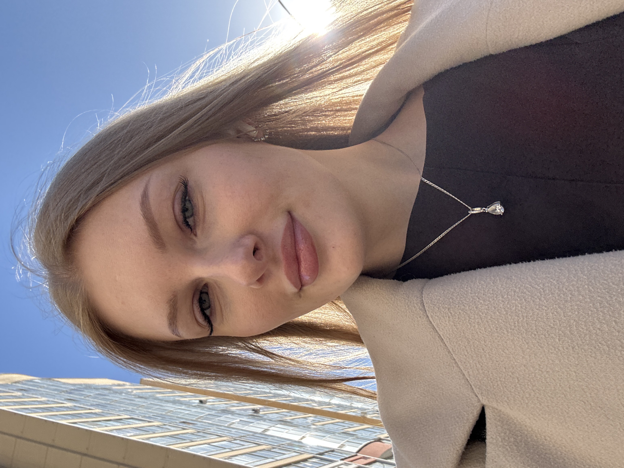 A young woman with blonde hair poses in front of a building with many windows. The sky is bright blue. The woman is wearing a dark-colored shirt and a light-colored coat. She has a silver necklace with a gemstone pendant. The location is Perm, Russia.