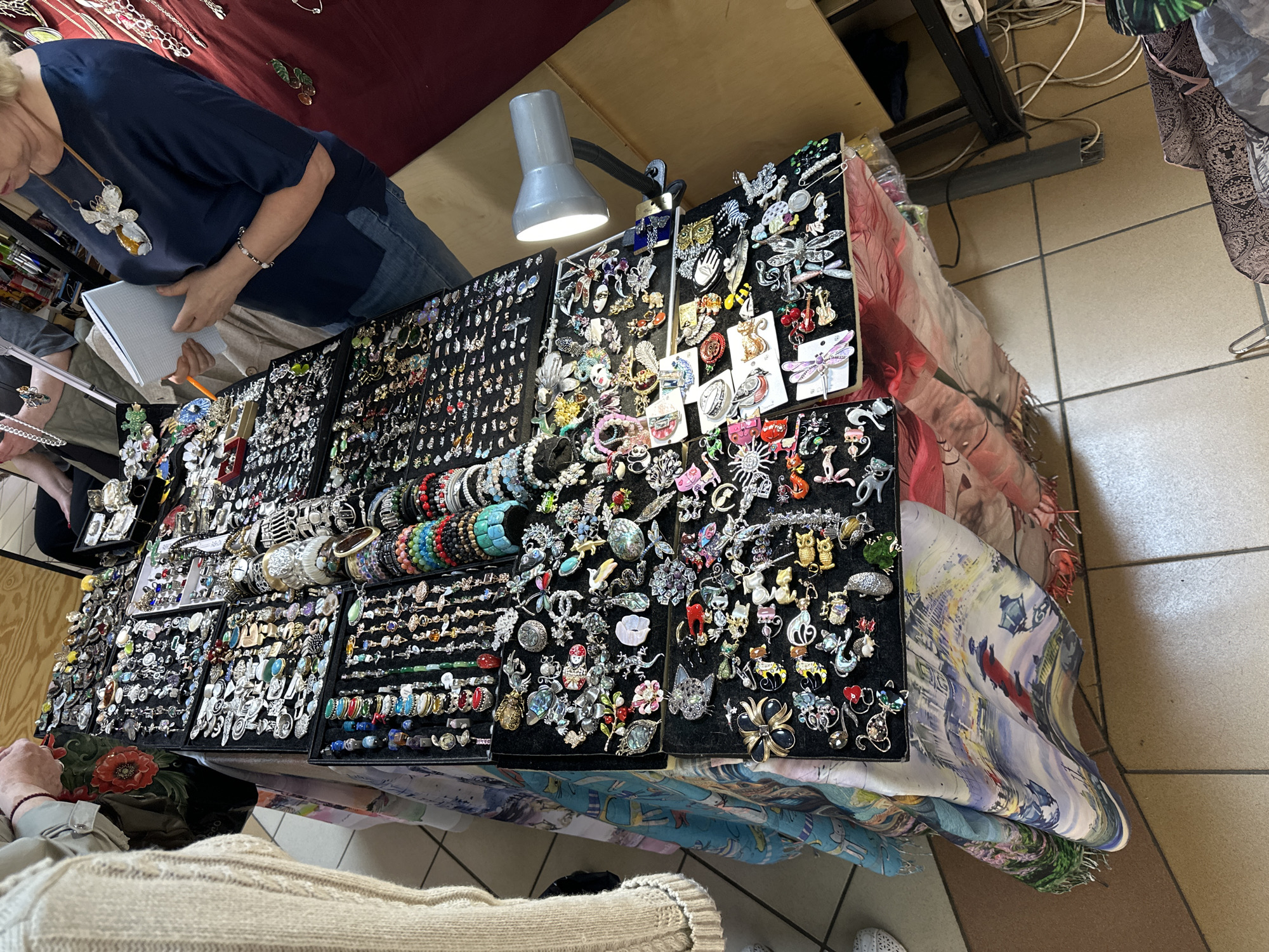A photograph captures a scene inside what appears to be a shop or market stall. On a table covered with patterned fabric, an extensive collection of jewelry, including rings, bracelets, and brooches, is displayed on black trays. A gooseneck lamp is positioned to illuminate the items. Two individuals are present, one examining the jewelry while holding a notepad and pen, and another with their back to the viewer. The backdrop consists of a red-toned fabric and a shelf with various items. The flooring is tiled. While there is no confirmation, based on the appearance of the items sold, the scene might be occurring in Saint Petersburg, Russia.