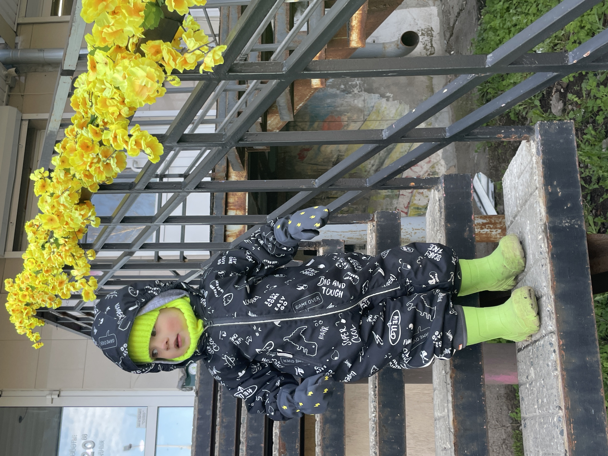 A young child, likely a toddler, is the central subject of this outdoor photograph taken in Izhevsk, Russia. The child is dressed warmly in a black, hooded winter overall patterned with white drawings and English text, a vibrant neon green beanie, and matching neon green rubber boots and gloves. They stand on a dark, weathered metal platform or step, looking slightly upward and to the left with a neutral, curious expression, their gloved hands gently raised.
The child is framed by a sturdy black metal structure, resembling a railing or fence, which extends diagonally across the image. Draped abundantly over this metalwork, especially to the child's left, are numerous artificial yellow flowers, adding a bright pop of color against the darker tones of the metal and the child's outfit.
The background on the left features a building with a reflective glass door, showing inverted Cyrillic text and reflections of an overcast sky. Behind the metal structure, glimpses of green vegetation, concrete elements, an industrial pipe, and a section of a painted wall or mural are visible, indicating an urban or semi-industrial outdoor setting.
The overcast sky suggests it is daytime, and the child's protective attire implies cool or cold weather.
Visible text on the child's overall includes phrases like "GAME OVER," "BIG AND TOUGH," "SUPER COOL," "ROARR," "HELLO," "SCARY FACE," "EASY ONE," "WILD LOVER," and "LUDER." On the reflected glass door, partial and inverted Cyrillic characters such as "ДНЫХ" and "аботы:", alongside numbers like "2088," are discernible.