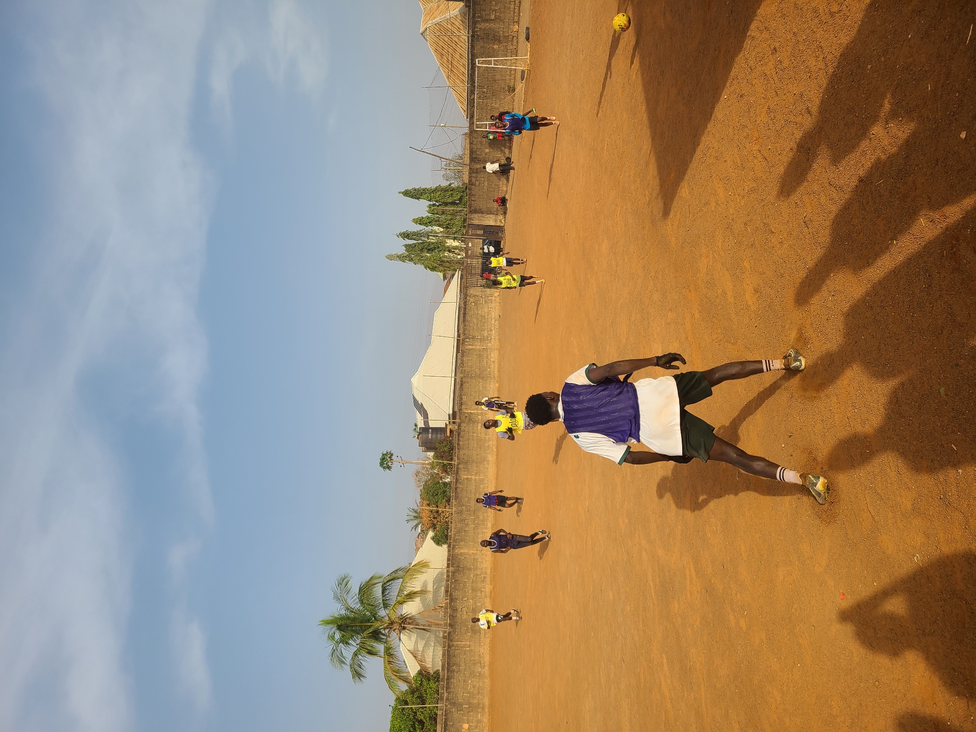 The media file presents an outdoor scene of a football (soccer) game played on a reddish-brown dirt field in Barakin Lerawi, Nigeria. The sky is bright and clear with minimal clouds, suggesting good weather conditions. In the background, a low brick wall separates the playing area from some green trees, including a prominent palm tree and several conical-shaped trees, and simple buildings with light-colored roofs. A white goal post with a net is visible at the far end of the field.
Several males, appearing to be young adults or teenagers, are scattered across the field, participating in or observing the game. In the immediate foreground, a player is captured from the mid-torso down, wearing a purple and white jersey, dark shorts, white socks, and green and yellow athletic shoes, facing away from the camera. Other players further back are wearing various colored jerseys, including yellow, blue, and white, indicating different teams or casual play. A yellow football is visible in the upper right portion of the frame, seemingly in mid-air or recently having landed.
The presence of long, distinct shadows cast by the players and structures suggests that the time of day is either early morning or late afternoon, with the sun low in the sky, creating a warm, golden illumination across the field. The overall scene depicts an active community sporting event. No discernible text is visible on any jerseys, signs, or objects in the image.