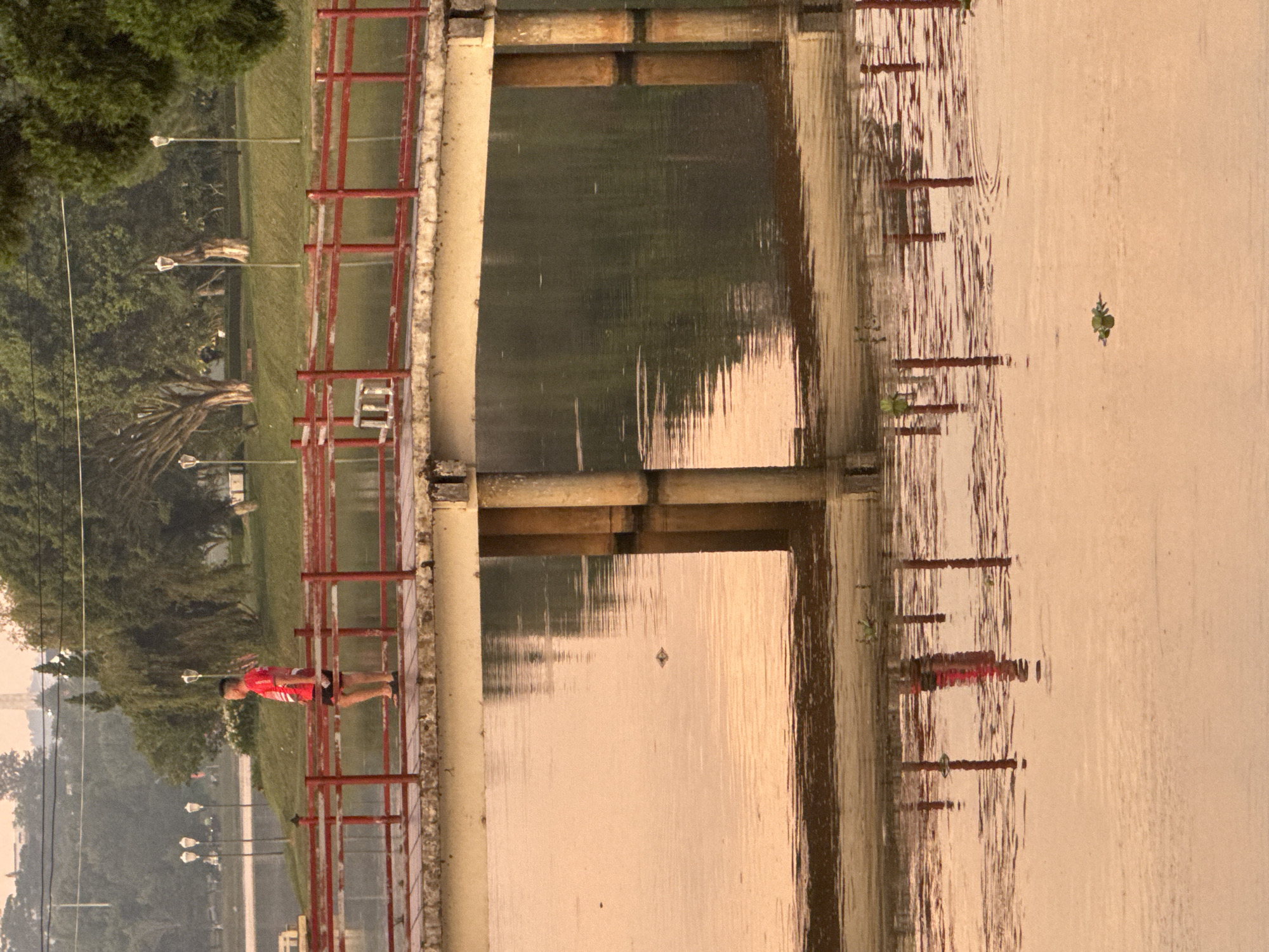 A person wearing a red shirt and black shorts stands on a wooden bridge with red railings over a body of water. The bridge has concrete pillars supporting it, and these pillars are reflected in the water. The water is calm and reflects the bridge and the surrounding trees. There are streetlights and trees visible in the background, suggesting a park or scenic area. The sky is overcast and the lighting suggests it is either early morning or late evening. This scene is consistent with locations such as the Xuan Huong Lake area in Phường Xuân Hương - Đà Lạt, Vietnam, known for its bridges and reflective water.