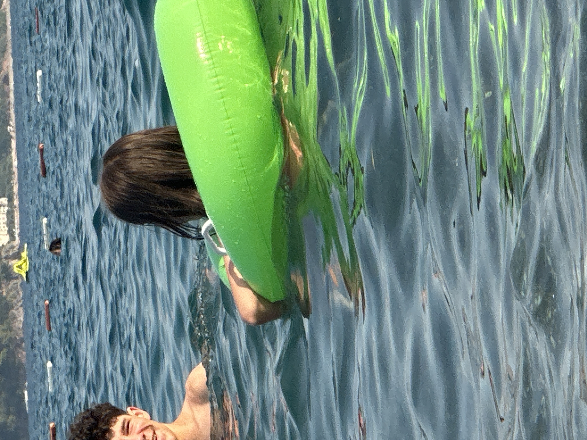 The image captures a scene in the Adriatic Sea off the coast of Boreti, Montenegro. In the foreground, two individuals are partially submerged in the water. On the left, a young male with dark, curly hair is smiling and looking towards the camera. He is bare-chested. To his right and slightly further back, another person, likely a young female, is seen from behind, her dark hair visible as she holds onto a bright green inflatable tube. The water is a deep blue with noticeable ripples, indicating a gentle wave motion. In the background, several buoys are visible, marking a swimming area. Further in the distance, the coastline of Boreti can be seen, characterized by lush green hills and some buildings scattered across them. The overall impression is of a relaxed summer day at the beach.